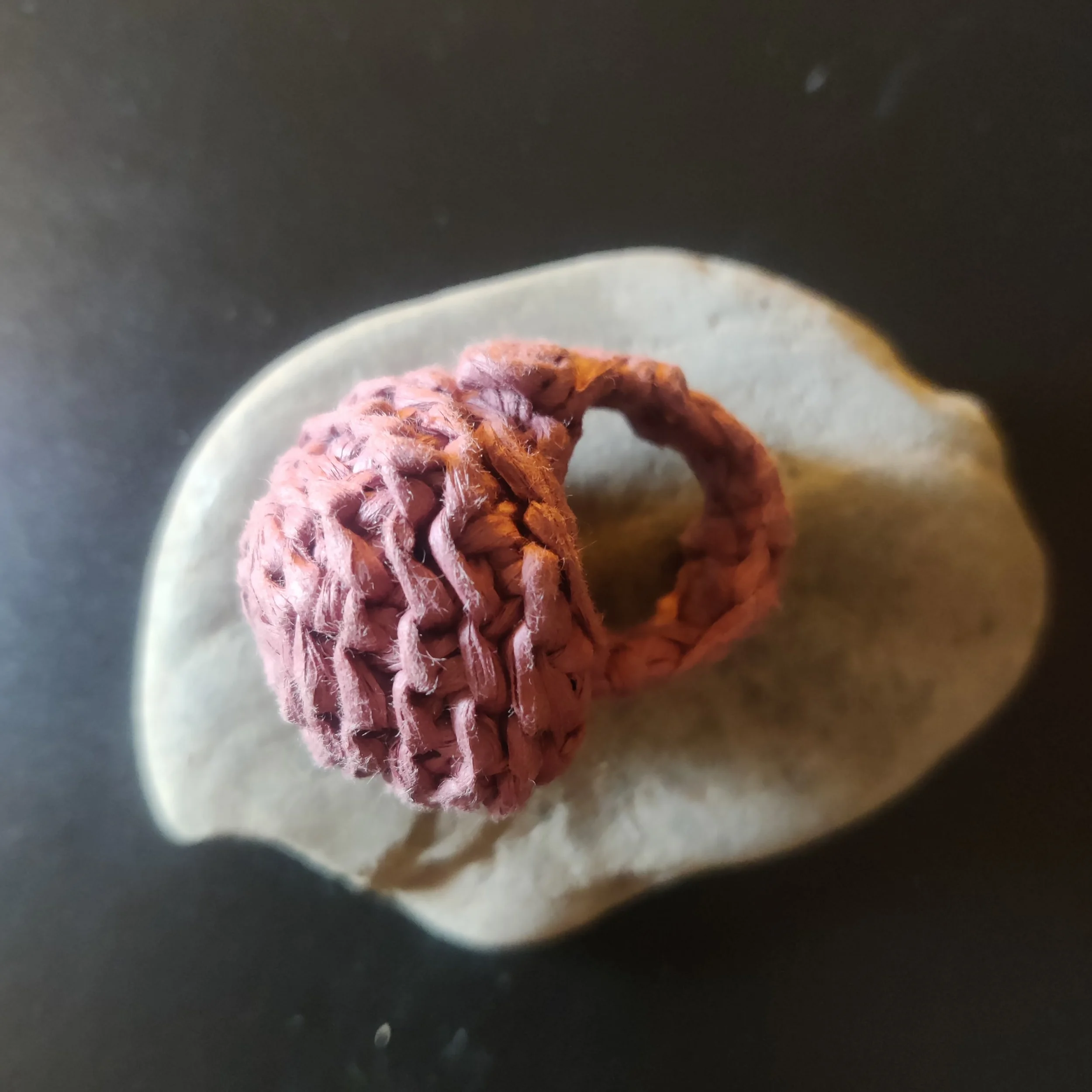 Anillo redondo de rafia rosa tejido en forma de espiral, colocado sobre una piedra blanca.