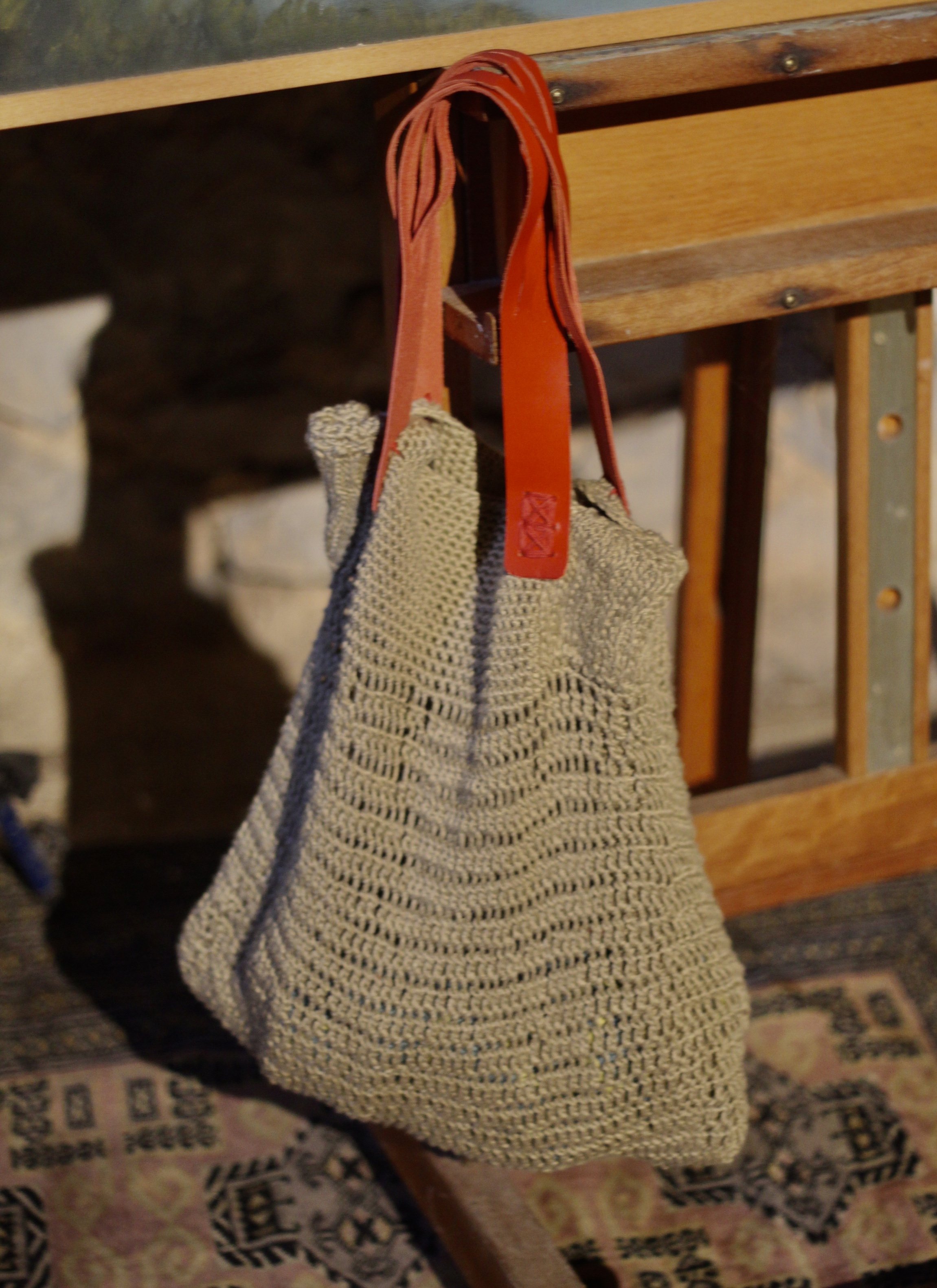 Bolsa tejida colgada de un respaldo de madera, con tirantes de cuero rojo, en un entorno interior.