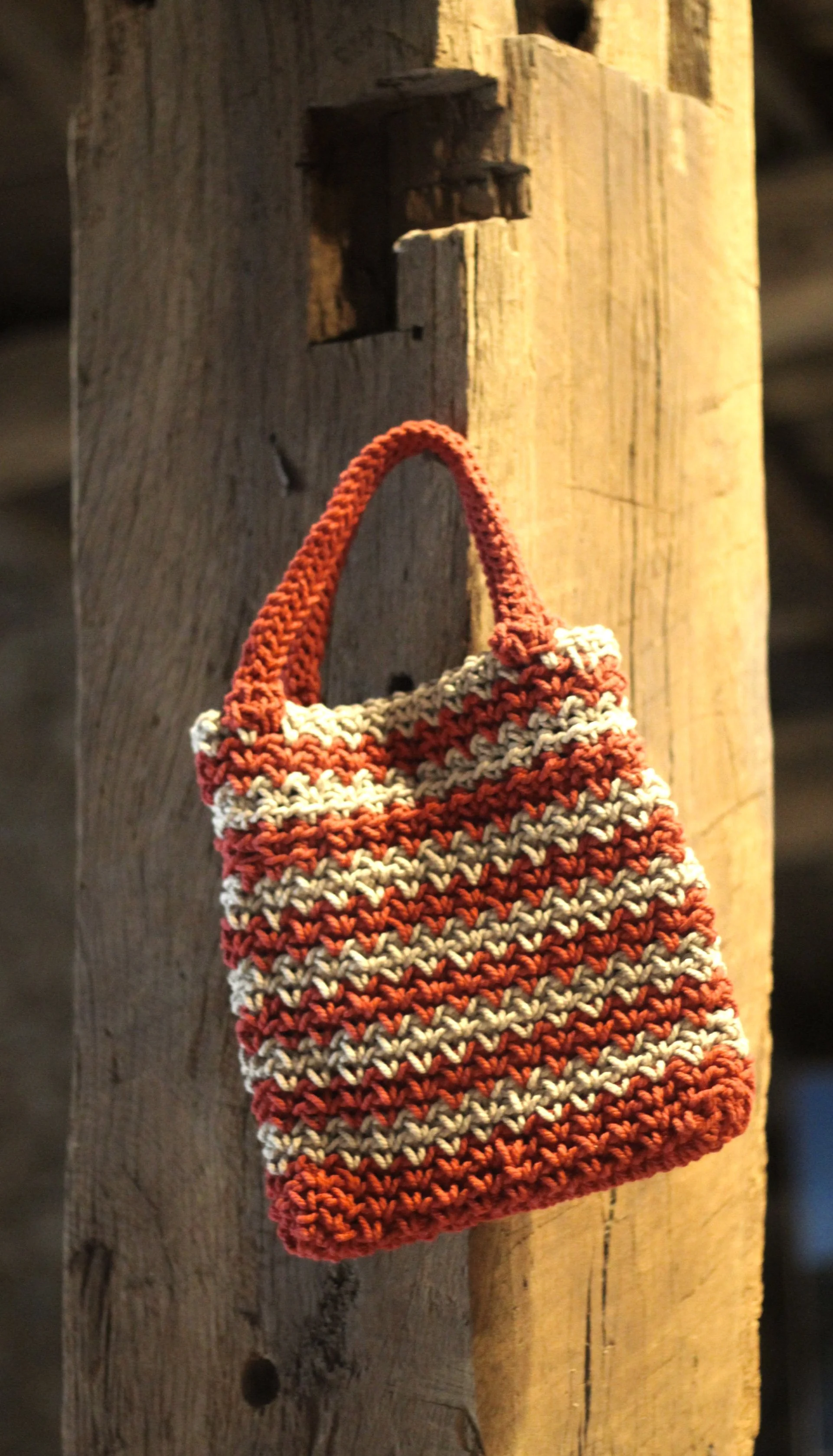 Un bolso tejido a crochet en algodón en rayas rojas y crudas, colgado en una estructura de madera.