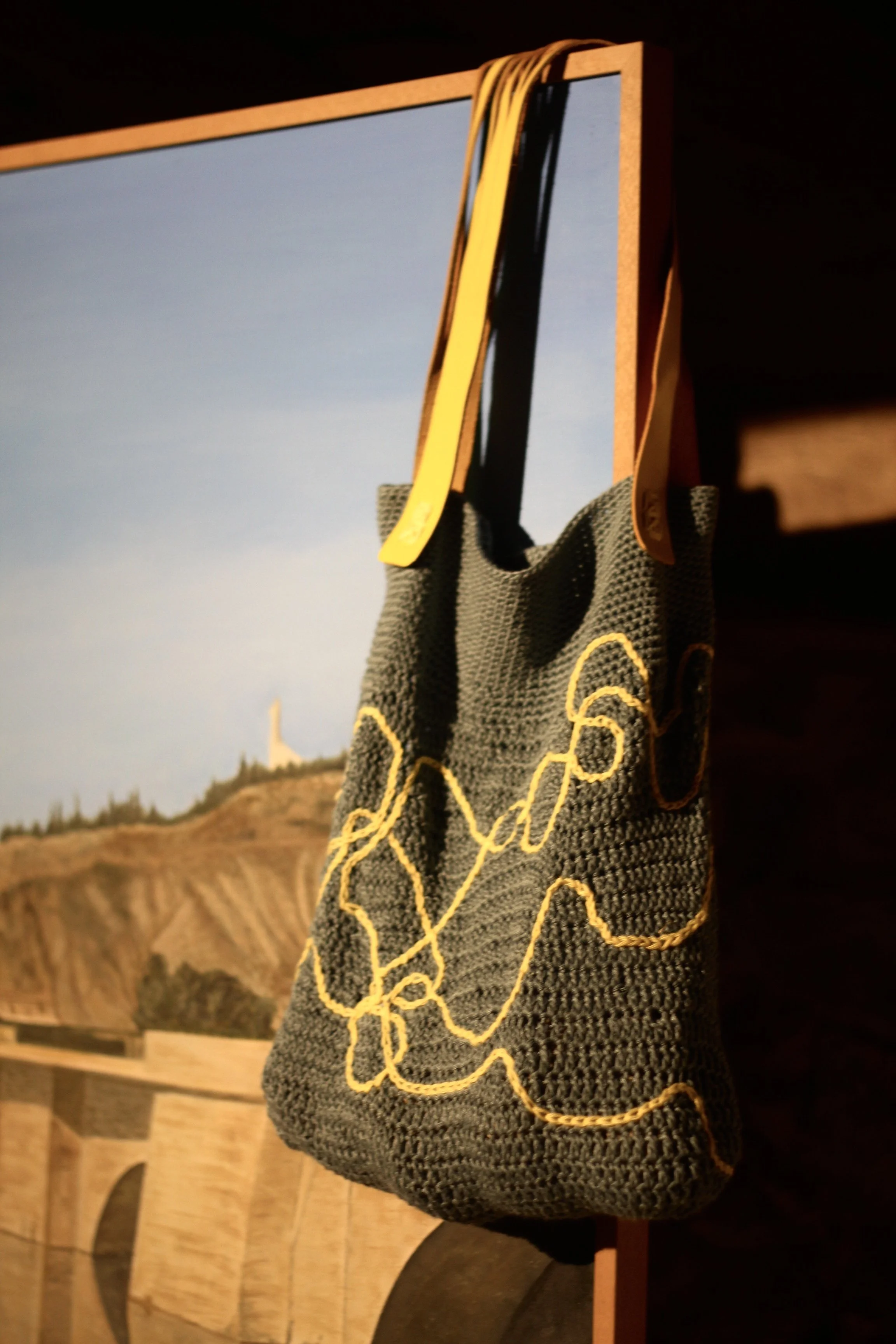 Bolso tejido en bambú azul con diseño de líneas de color amarillo, colgado de un marco de madera. En el fondo, una fotografía de paisaje con un puente y una colina.