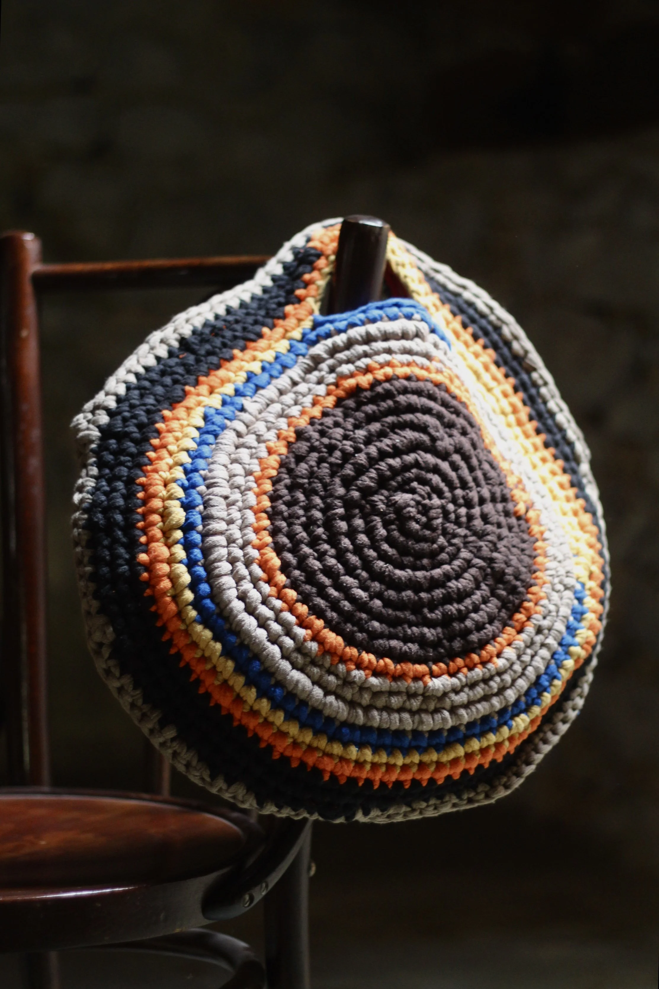 Un bolso tejido de forma circular con hilos de colores naranja, azul, gris, negro y blanco, colgado de la espalda de una silla de madera.