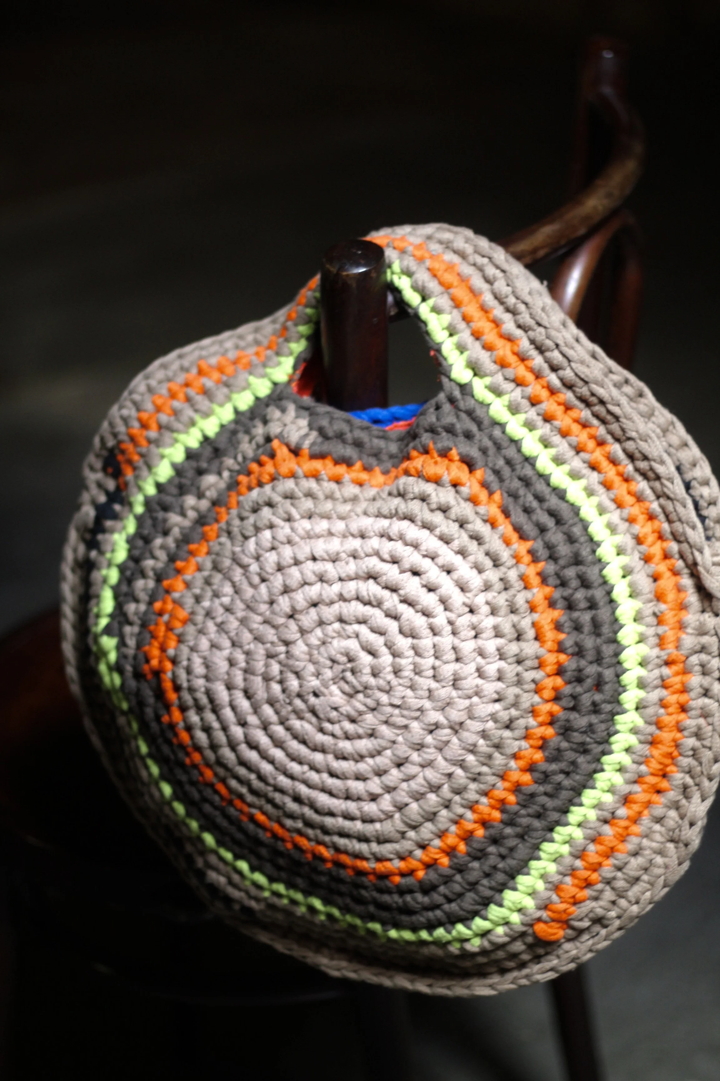 Bolso tejido de color gris con detalles en verde, naranja y azul, colgado de una silla de madera.