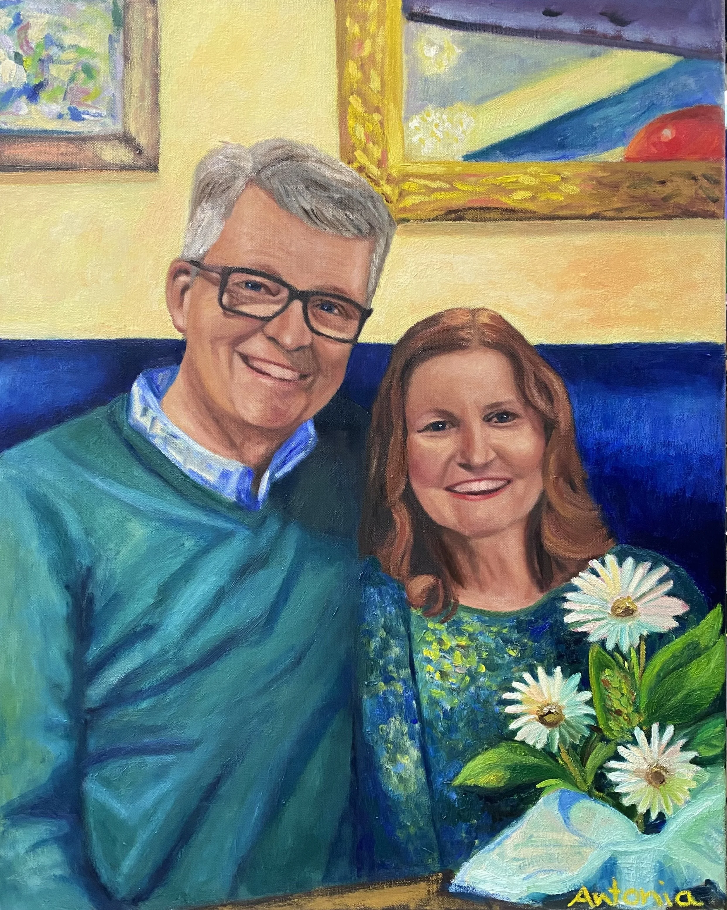 A painted portrait of a smiling man with gray hair and glasses, and a smiling woman with reddish-brown hair holding a bouquet of daisies. They are seated indoors with colorful framed artwork hanging on the yellow wall behind them.
