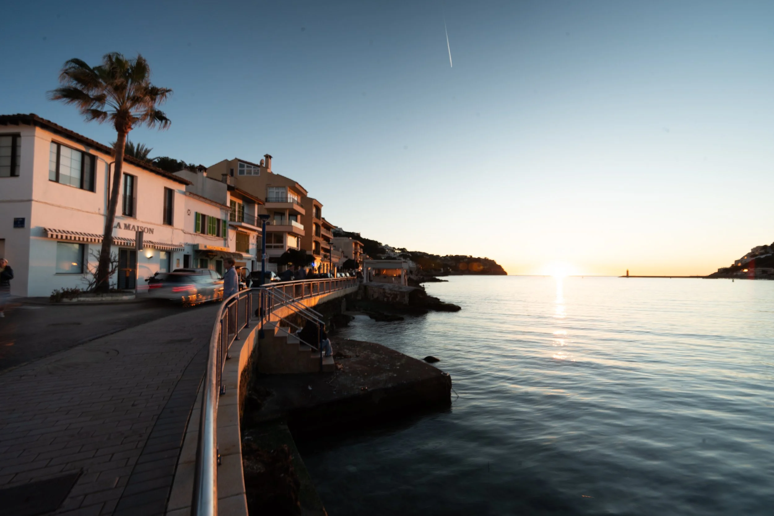 Sunset over a coastal town with buildings and palm trees, a promenade with a railing along the water, and the calm sea reflecting the sunset.