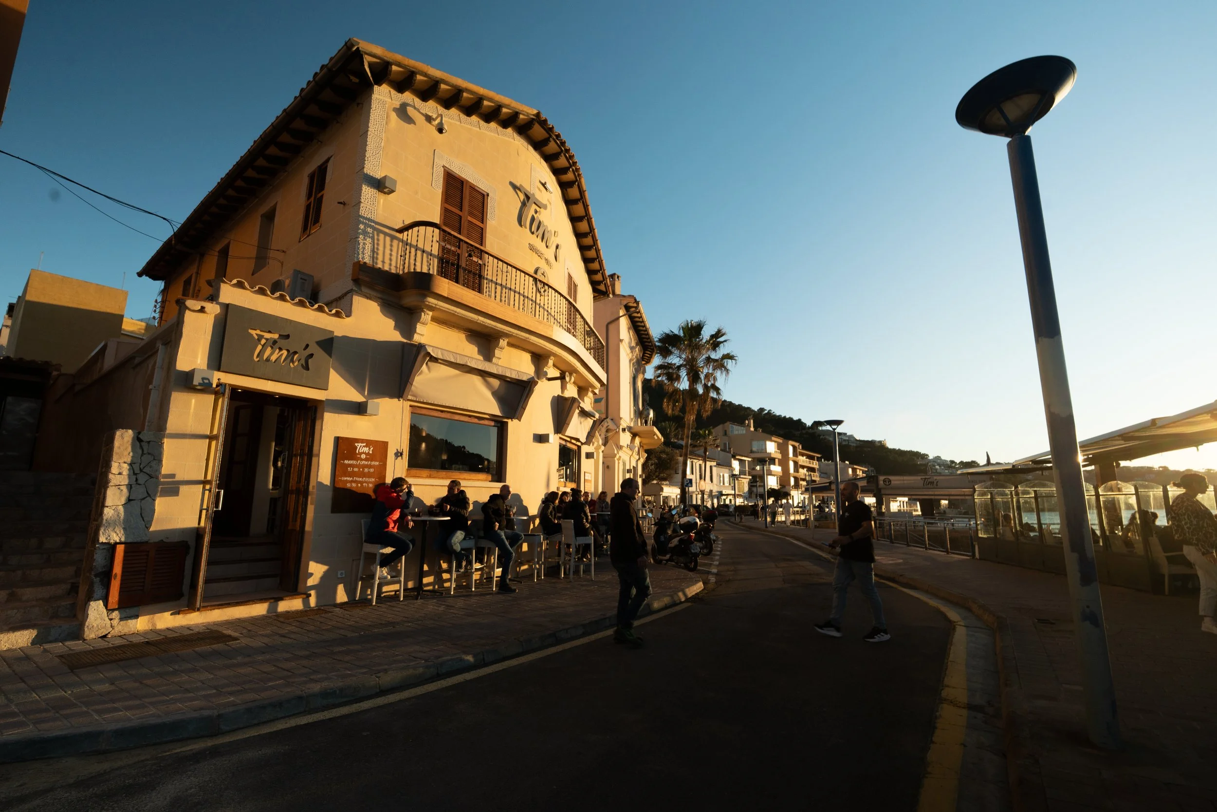 People sitting outside a restaurant called Timi's, with a street, palm trees, and hillside buildings in the background at sunset.