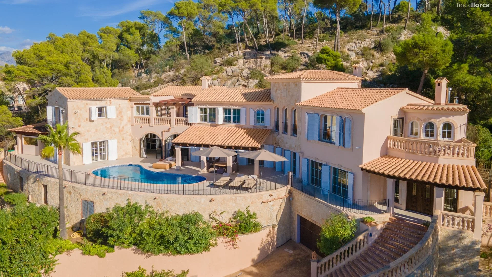 A large Mediterranean-style house with a swimming pool, surrounded by a stone wall fence and lush greenery on a hillside. The house has multiple levels, terracotta roof tiles, and arched windows with shutters. There are outdoor lounge chairs and umbrellas near the pool, with a curved staircase leading up to the main entrance.