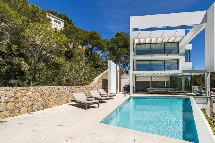 Modern white house with large glass windows, outdoor swimming pool, and lounge chairs on patio, against a backdrop of trees and blue sky.