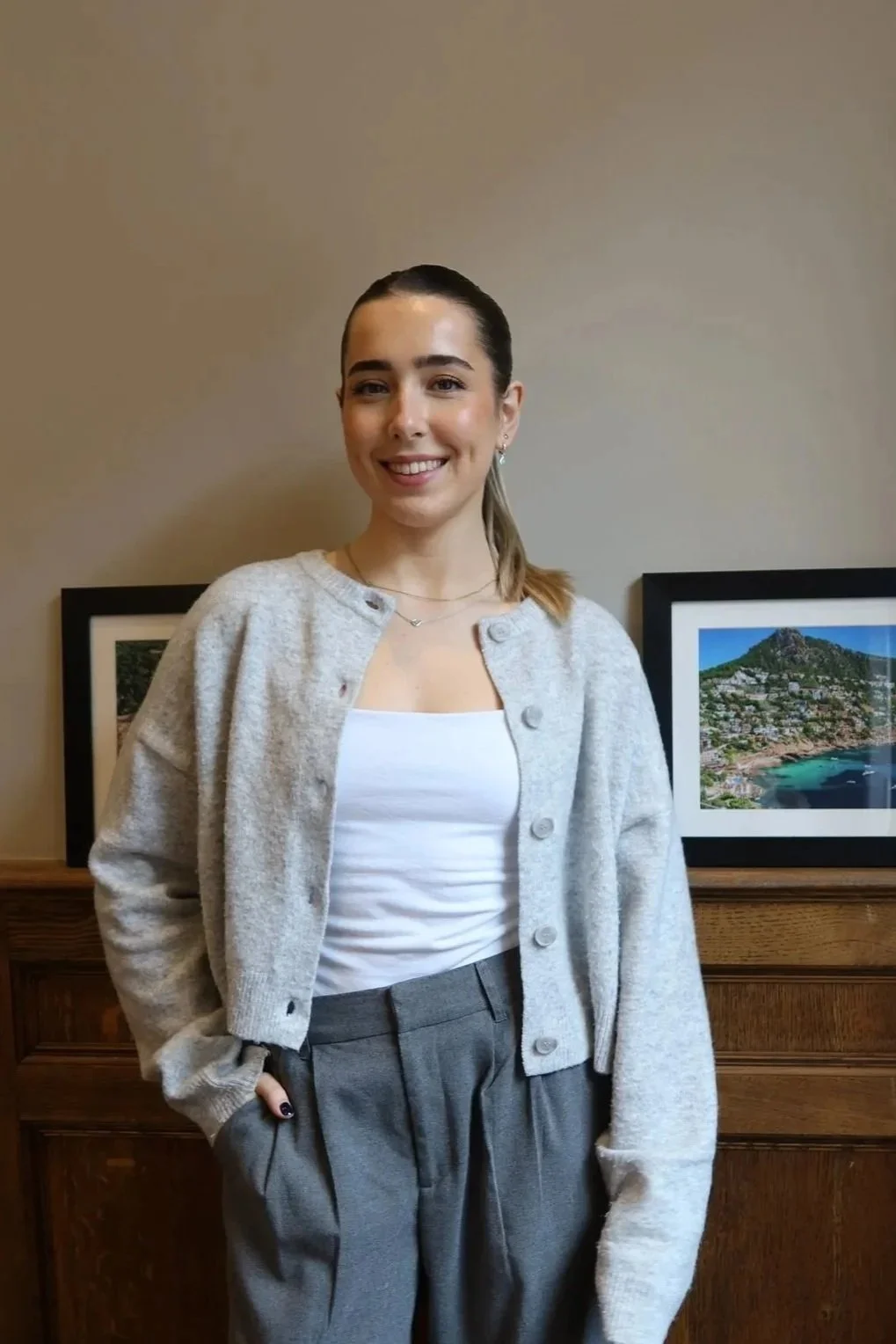 A young woman with brown hair tied in a ponytail, smiling, standing indoors in front of framed pictures of a landscape with a mountain and ocean, wearing a light gray cardigan over a white top and gray trousers.