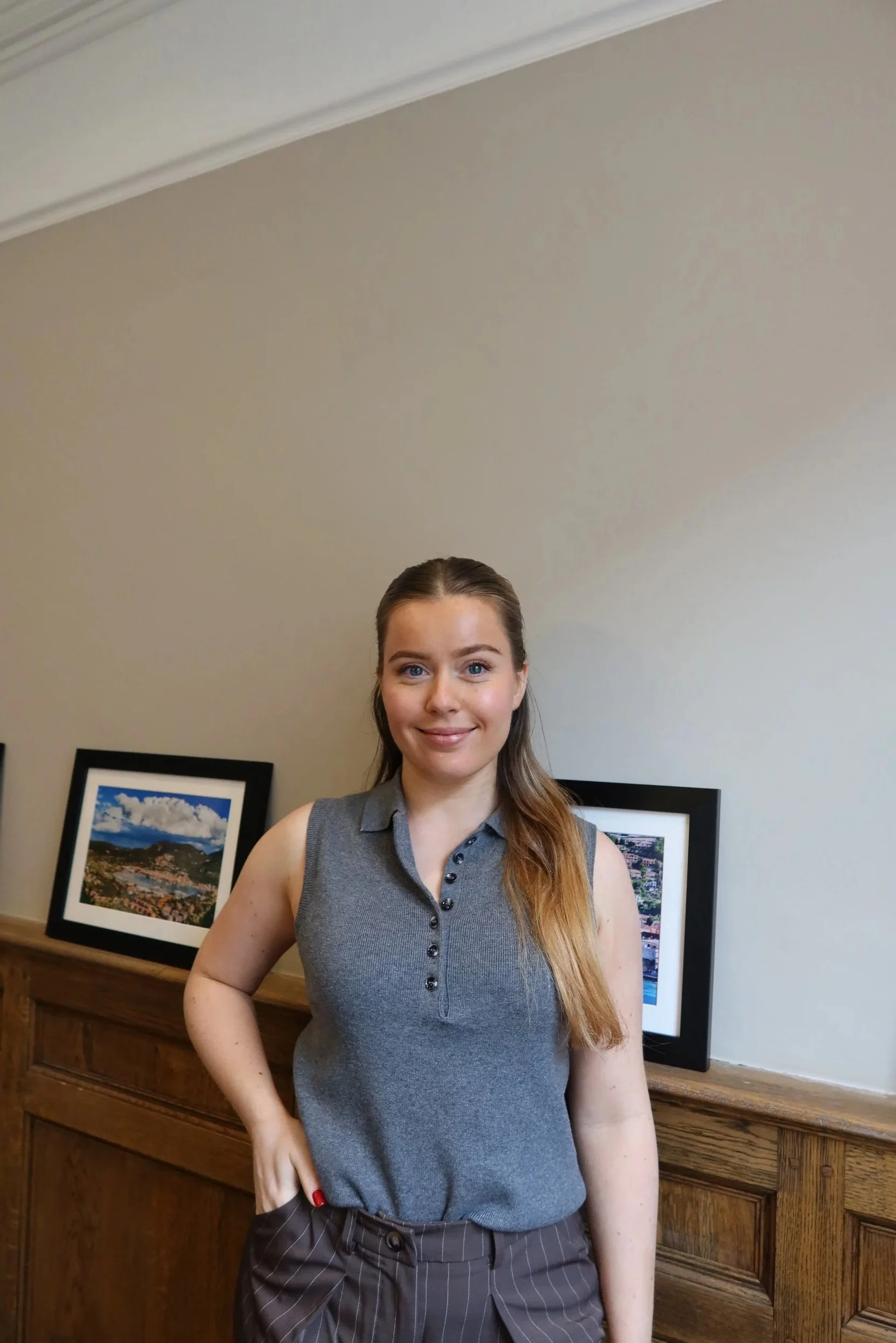 Young woman with long hair, wearing a sleeveless gray top and striped pants, standing indoors with framed photos on a wooden ledge behind her.