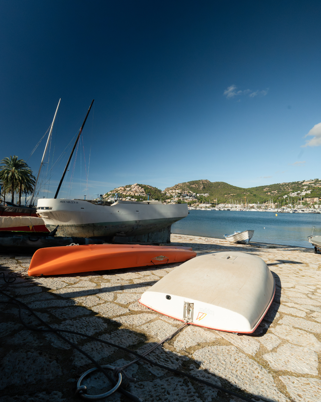 Boats on the shore of a marina with hills and a town in the background under a clear blue sky.