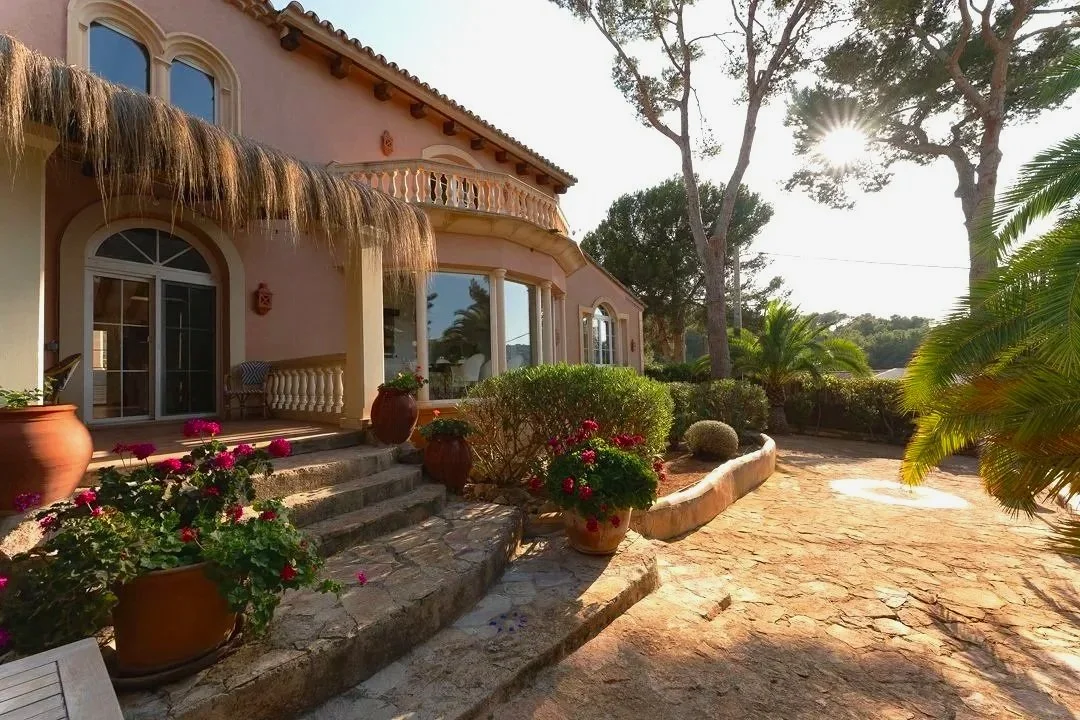 A house with a pink exterior, arched windows, and a balcony, surrounded by potted plants and flowers, with a stone pathway and trees nearby, under a bright sky with sunlight.