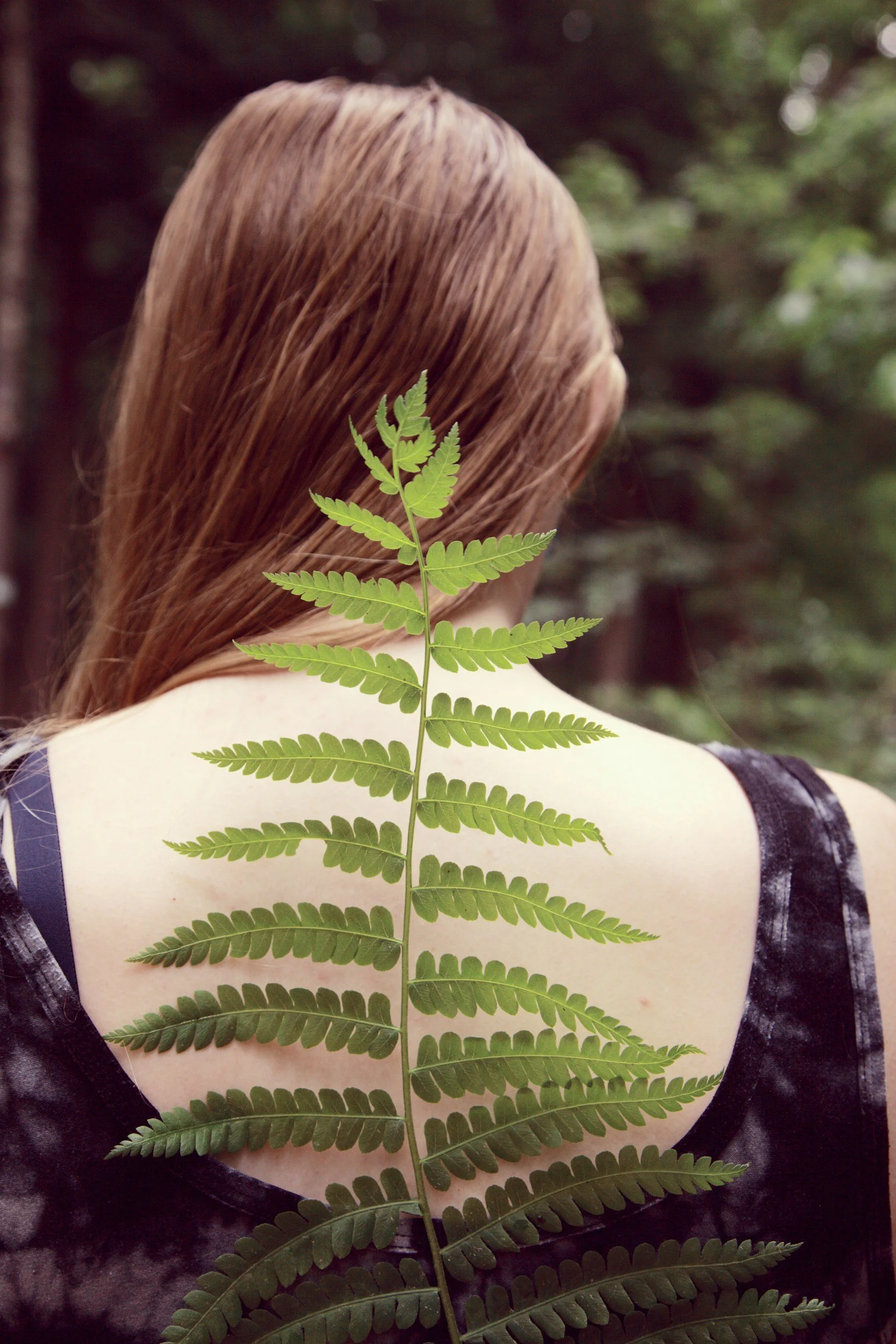 Back of a woman with long, light brown hair, wearing a dark, patterned sleeveless top, with a green fern leaf draping down her back. The background is a blurred green forest.