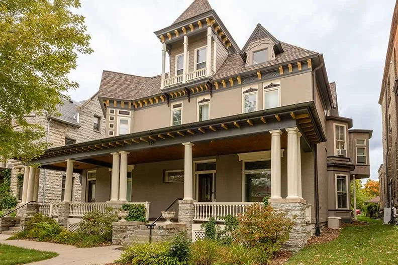 A large, Victorian-style house with a prominent front porch supported by white columns, multiple windows with decorative trims, and a turret at the top. The house is surrounded by a well-maintained garden and neighboring homes.
