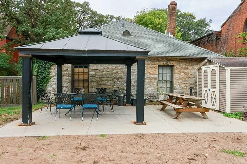 Backyard patio with a covered seating area featuring a metal table and six chairs, a wooden picnic table, a small storage shed, and a stone house in the background.