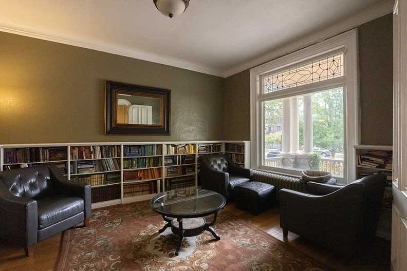 Living room with a large window, bookshelf, two black armchairs, one black loveseat, a round glass coffee table, a mirror on the green wall, and a patterned rug.