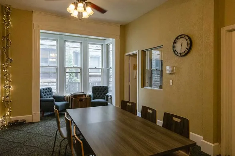 A dining room with a wooden table, six chairs, and a sitting area with two armchairs and a small table near large windows. There is a ceiling fan with lights, a clock on the wall, and string lights on the side.