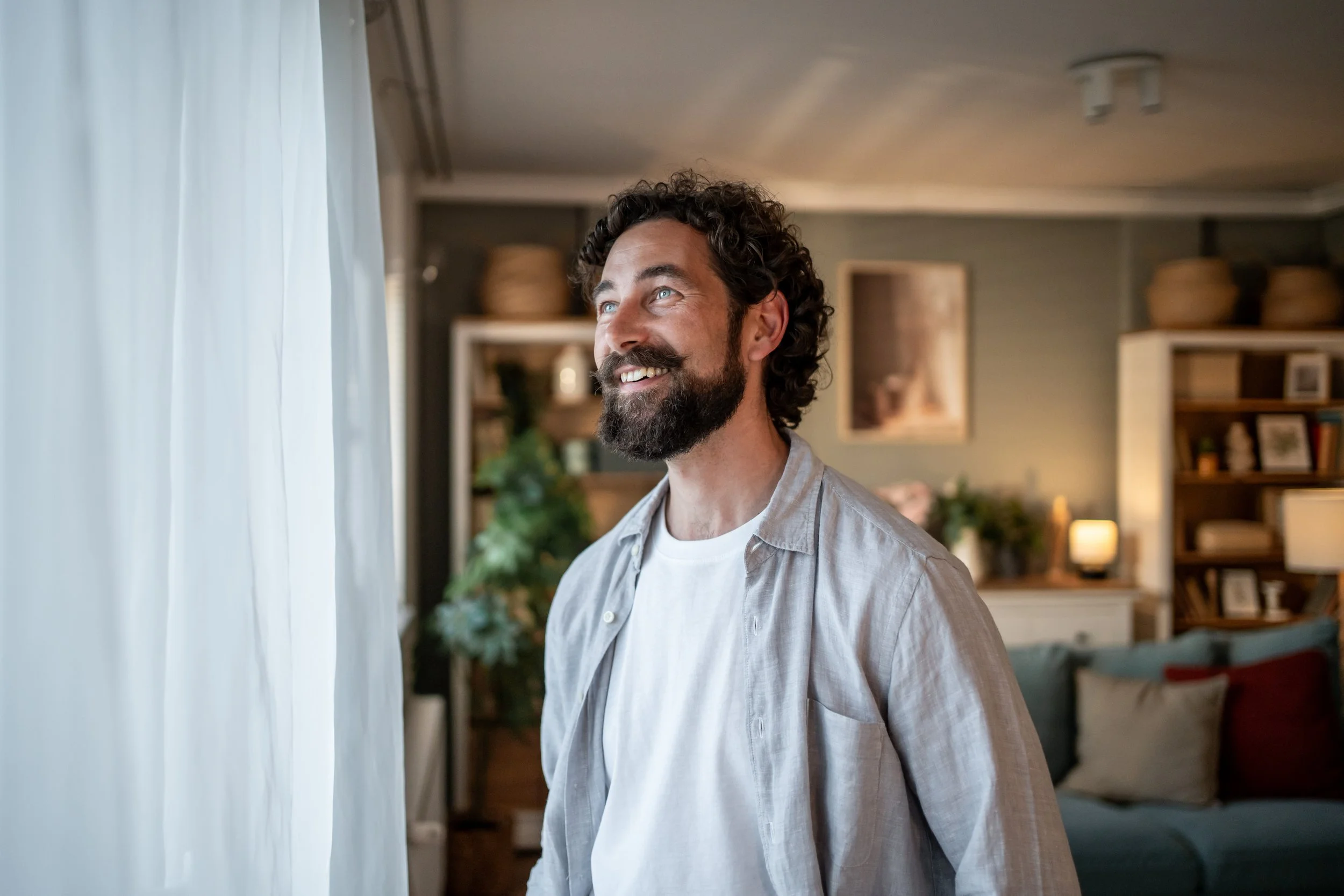 A man with curly hair and a beard smiling while standing by a window with white curtains in a cozy living room.
