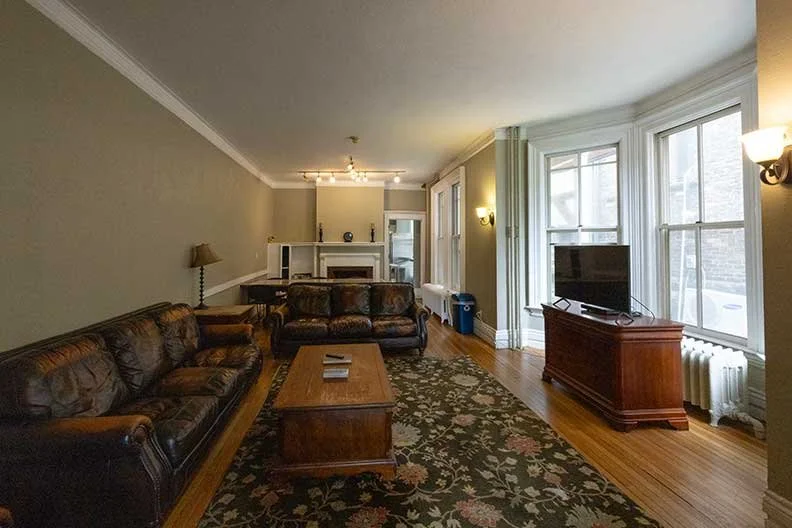 Living room with two leather sofas, a coffee table, a patterned rug, a TV on a wooden stand, large windows, and wall-mounted lamps.