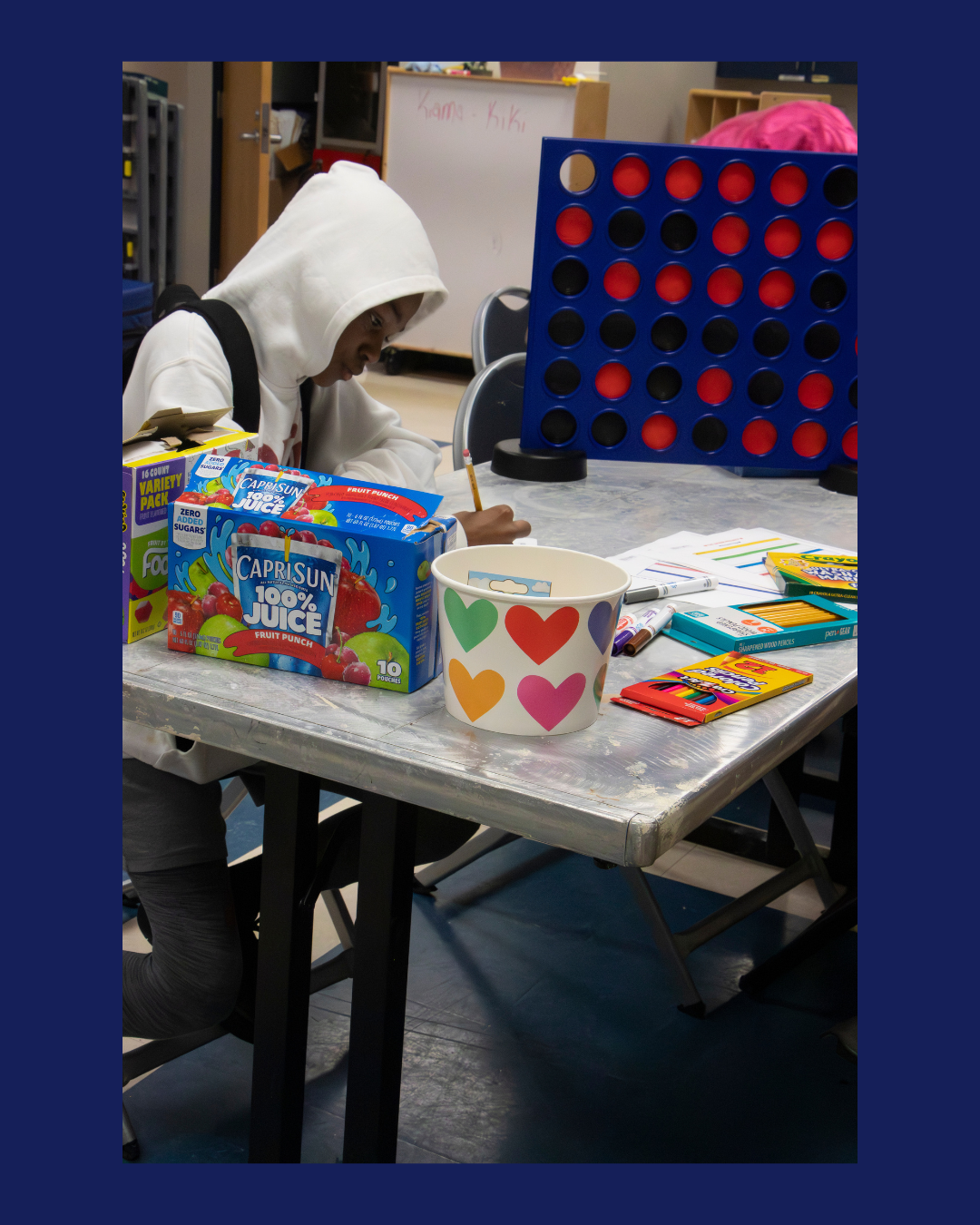 A young boy wearing a white hoodie playing Connect Four at a table with juice boxes, a heart-patterned cup, and drawing supplies.
