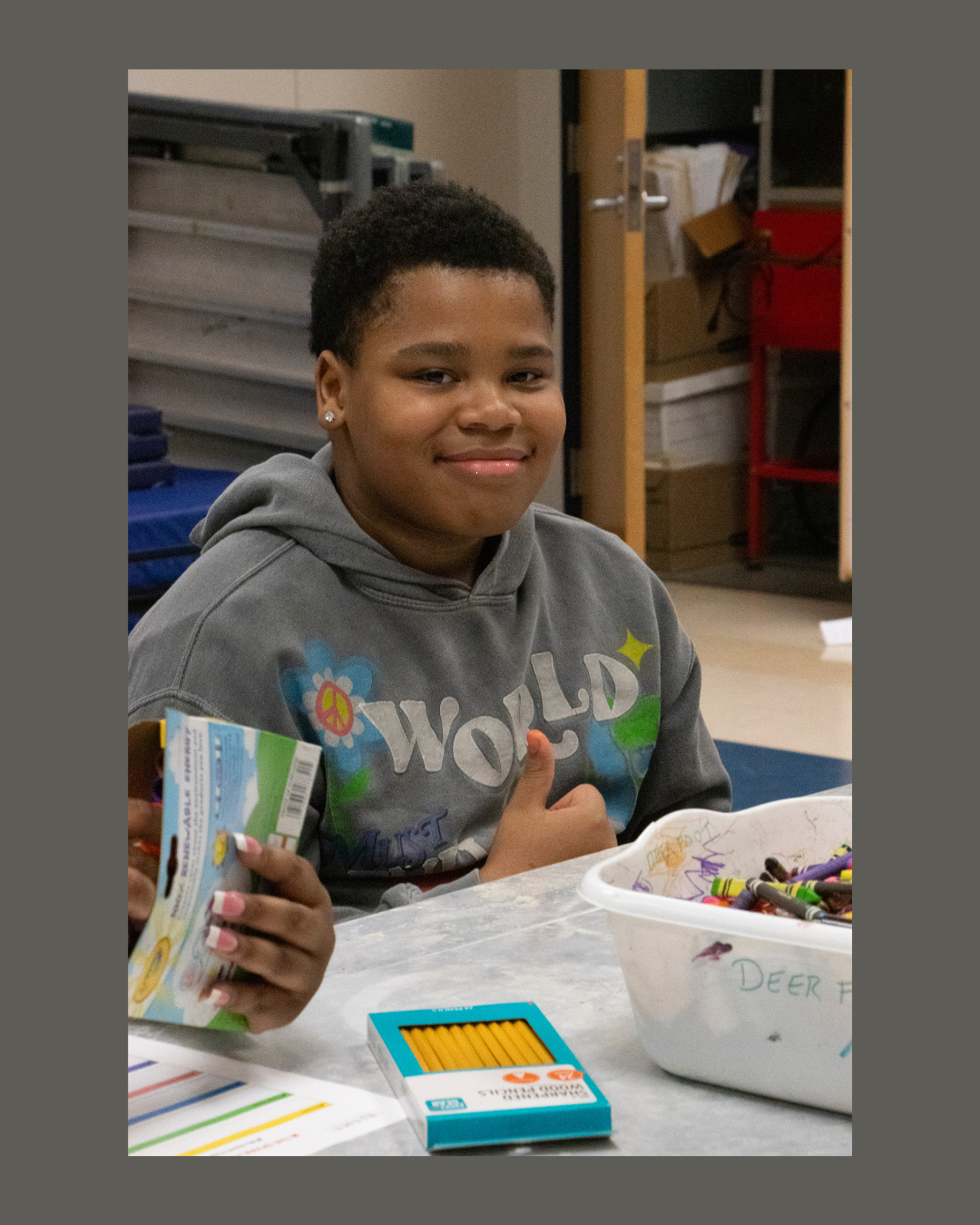 A child sitting at a table with school supplies, giving a thumbs up. He is wearing a gray hoodie with colorful designs.