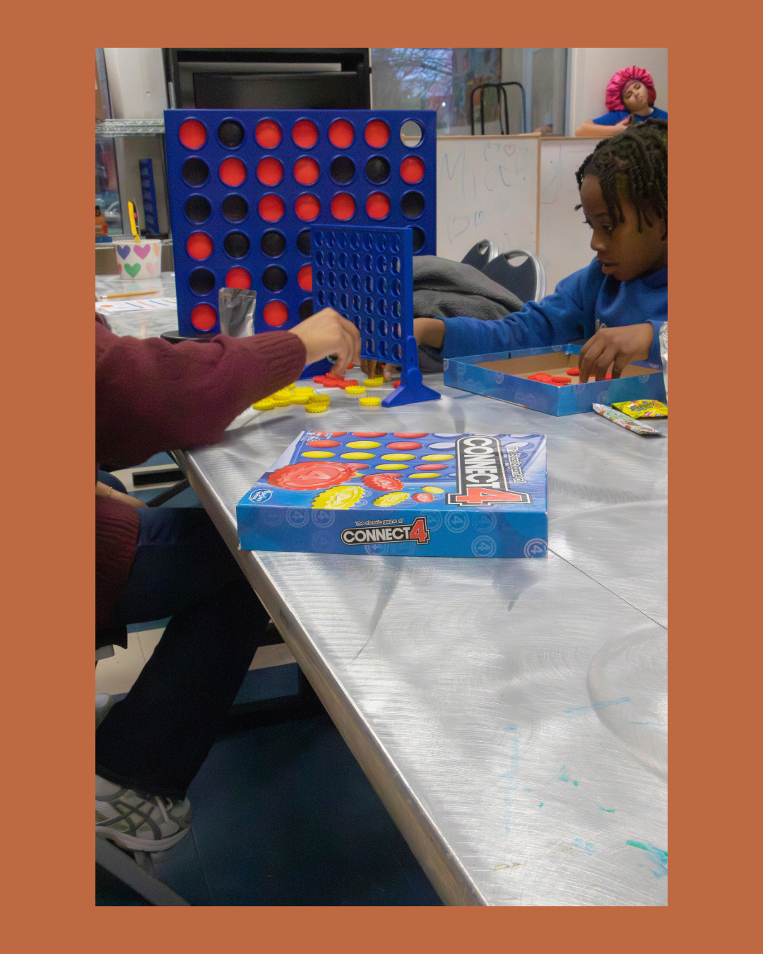 Two children playing Connect 4 at a silver table indoors, with a whiteboard, chairs, and a window in the background.