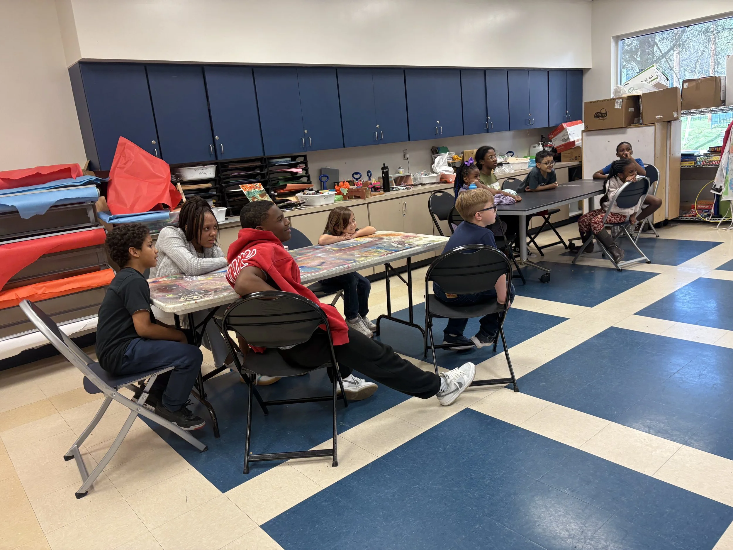 A group of children sitting and watching something in a classroom with blue cabinets, a table with art supplies, and shelves filled with boxes and books.