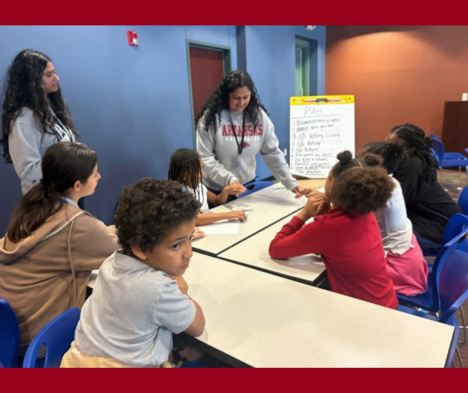 A group of children seated around a table in a classroom, listening to a woman standing and pointing at a whiteboard with notes. The classroom has blue walls and a door.