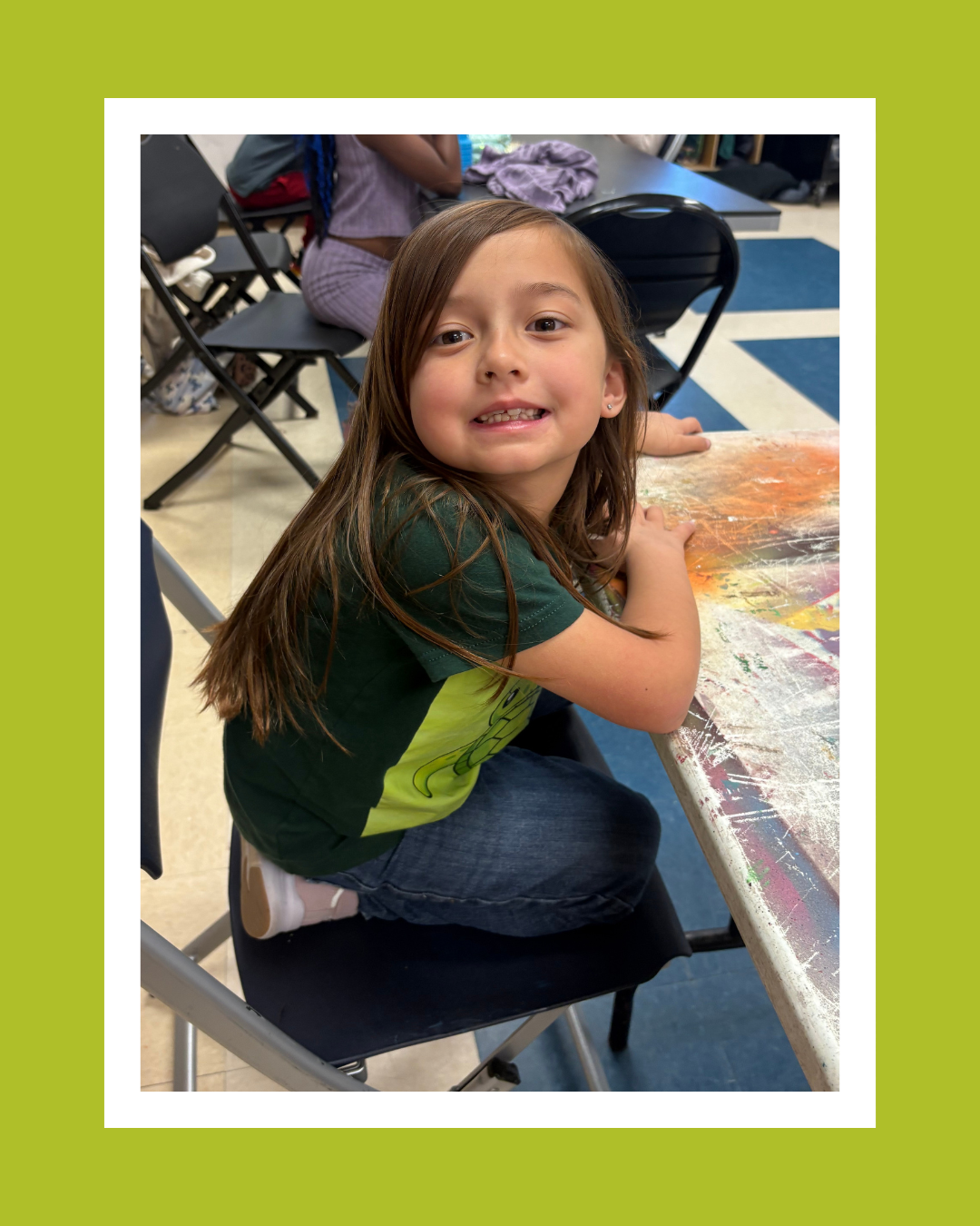 A young girl with long brown hair sitting at a paint-splattered table in a classroom, smiling at the camera.