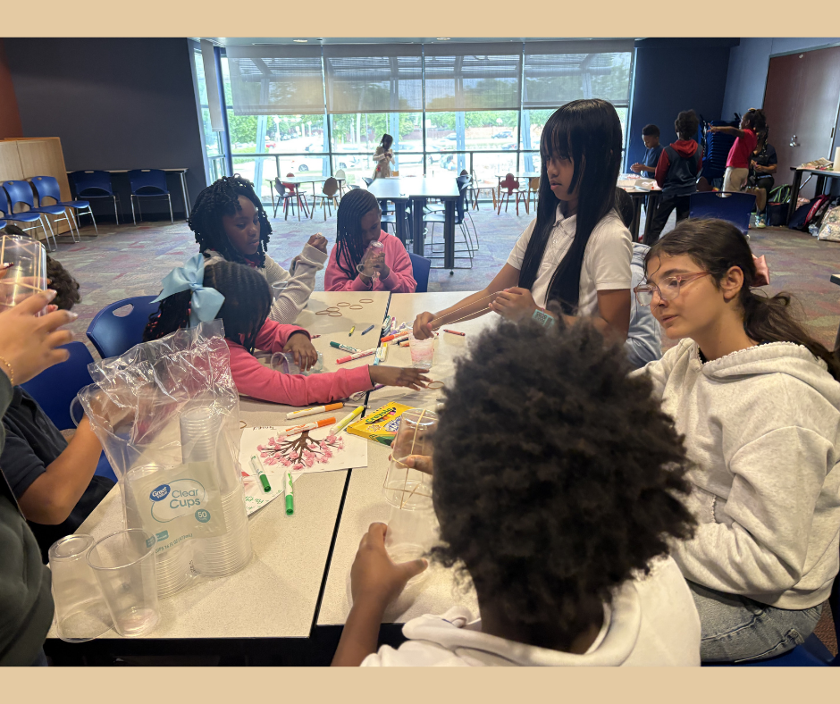 Children sitting around a table engaged in arts and crafts, with some using markers, paper, and other supplies, in a well-lit room with large windows.
