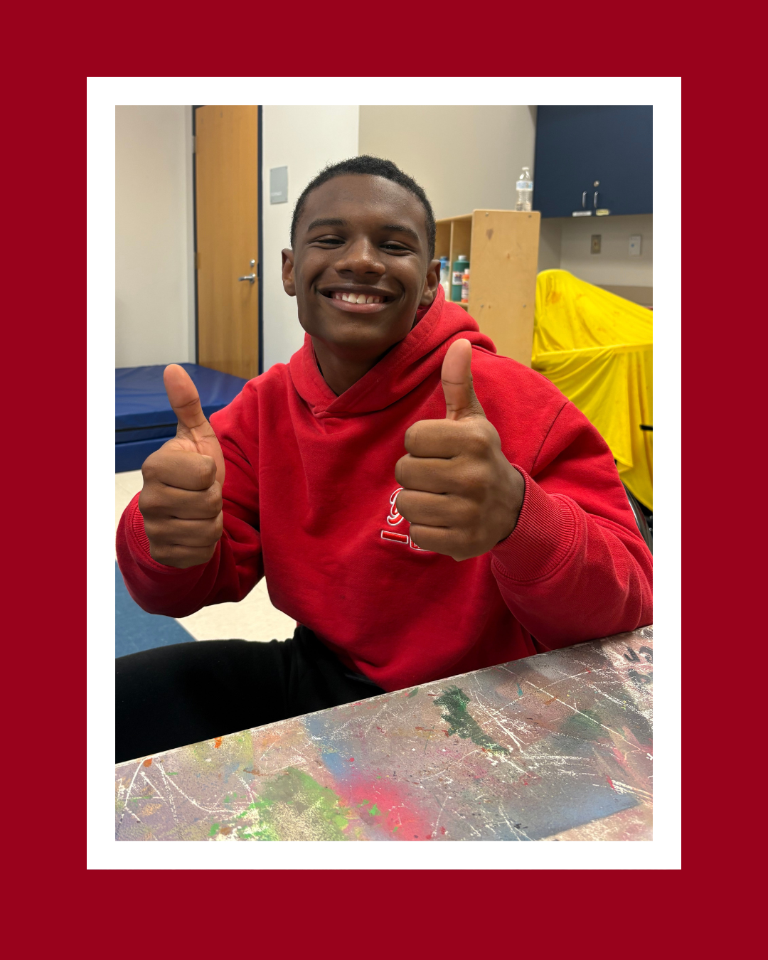 Young man in red hoodie giving two thumbs up and smiling, sitting at a table with colorful paint marks, in a room with a door, a shelf, and covered items in the background.