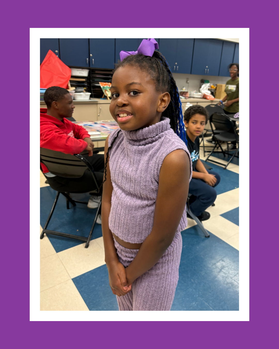 A young girl in a purple sleeveless knitted top and pants, with a purple bow in her hair, smiling at the camera with her hands clasped in front of her. Behind her are other children seated at tables in a classroom or activity room.