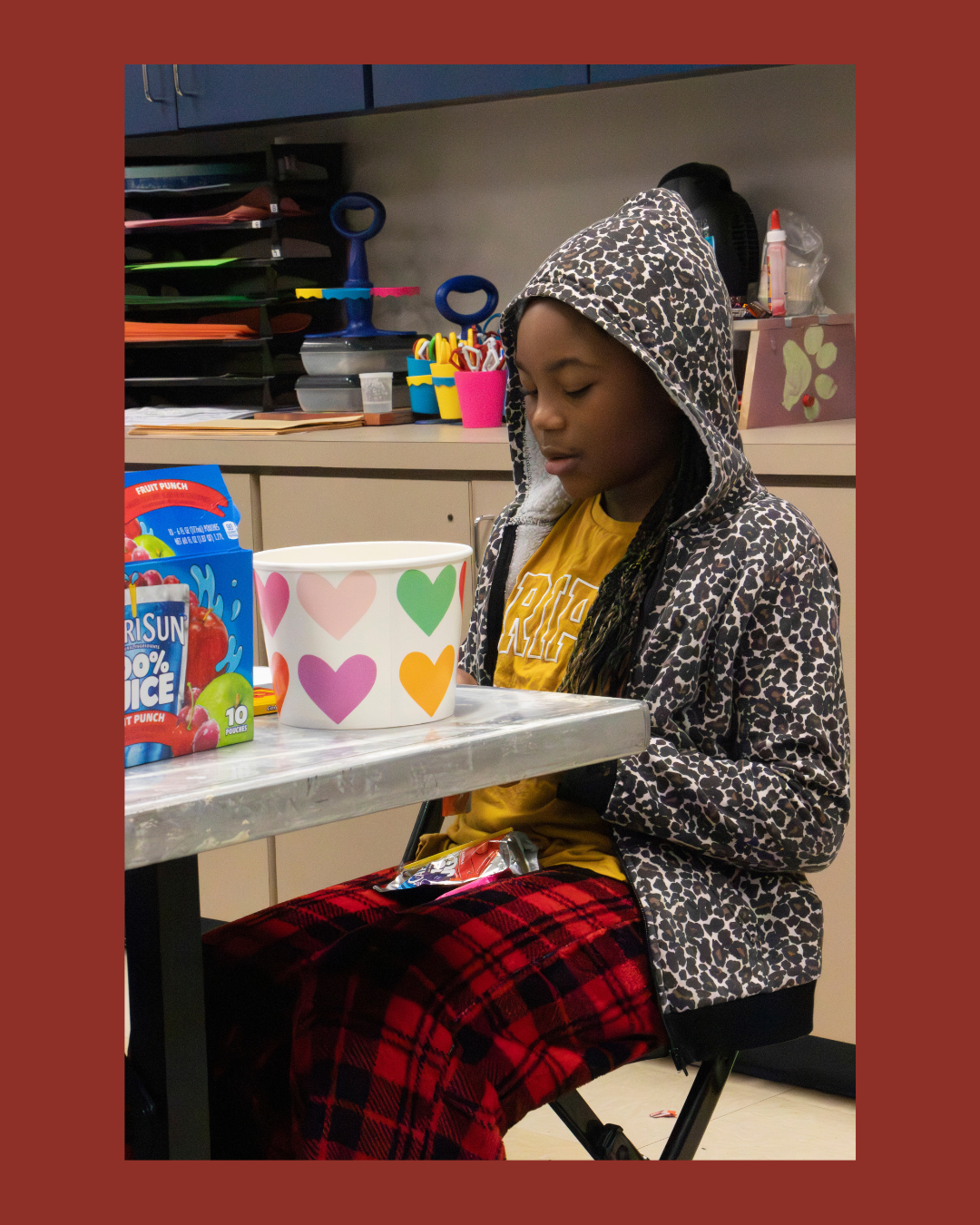 A young girl with a leopard print hoodie, yellow shirt, and red plaid pants sitting at a table with a colorful cup, snack, and juice box. She is looking down with her eyes closed, in a classroom or activity room setting with craft supplies in the bac