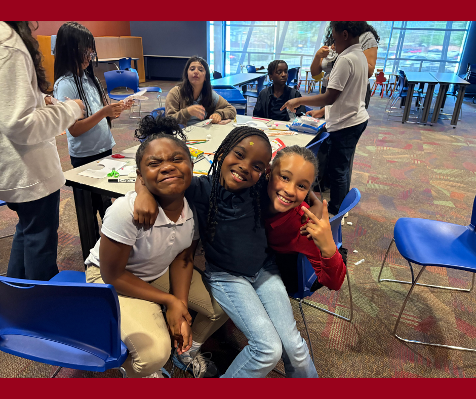 Three smiling girls sitting at a table in a classroom, with other children in the background engaged in activities.
