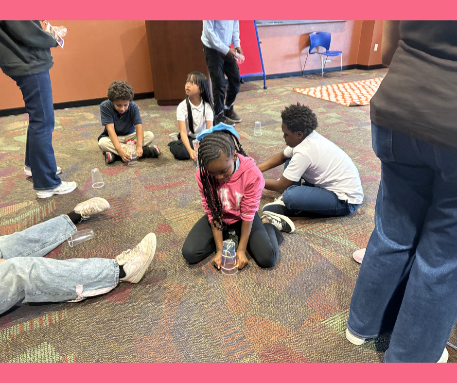 Children participating in a game or activity involving upside-down plastic cups on a carpeted floor in a room with orange and brown walls.