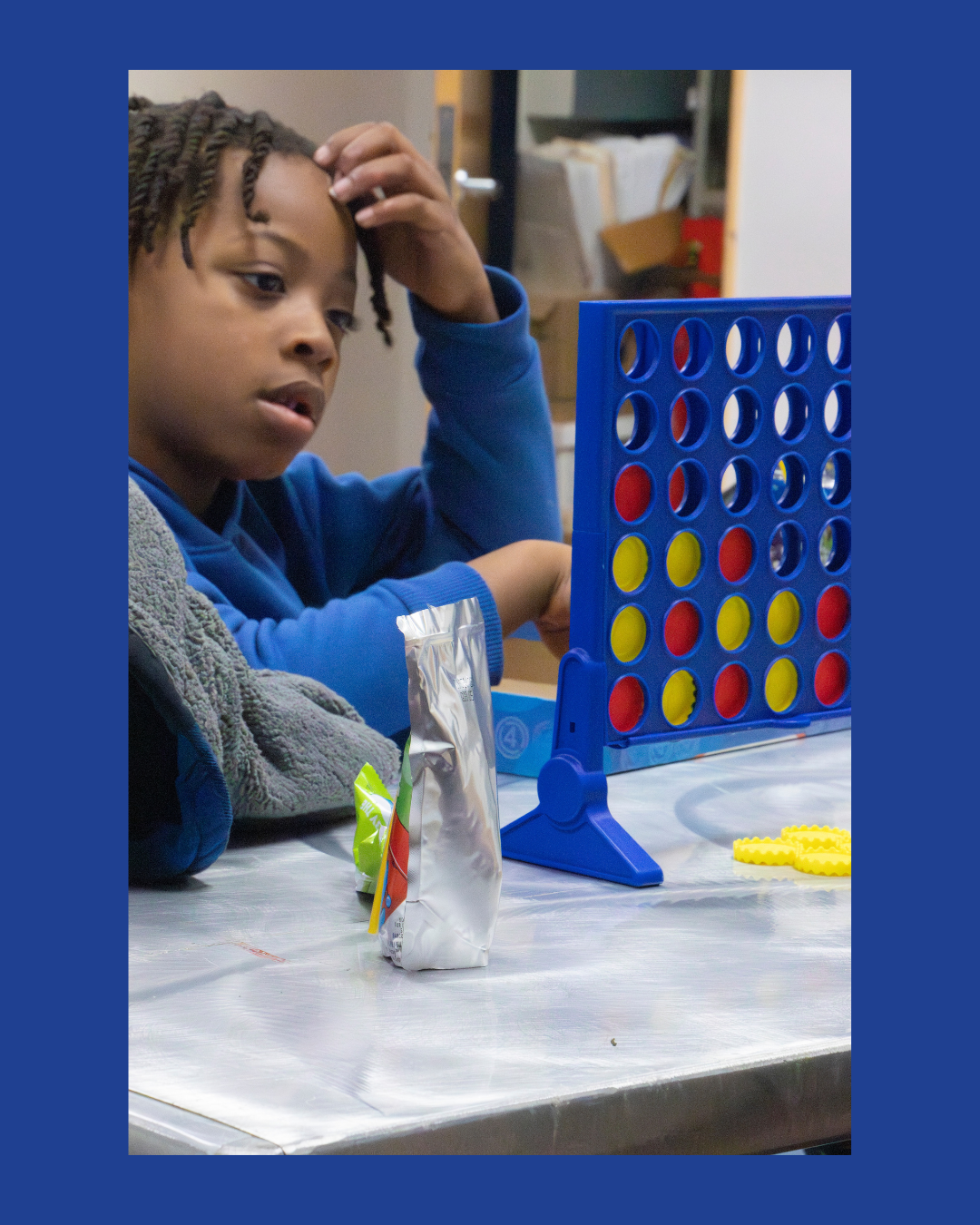 A young boy playing Connect Four at a table in a room with shelves and papers in the background.