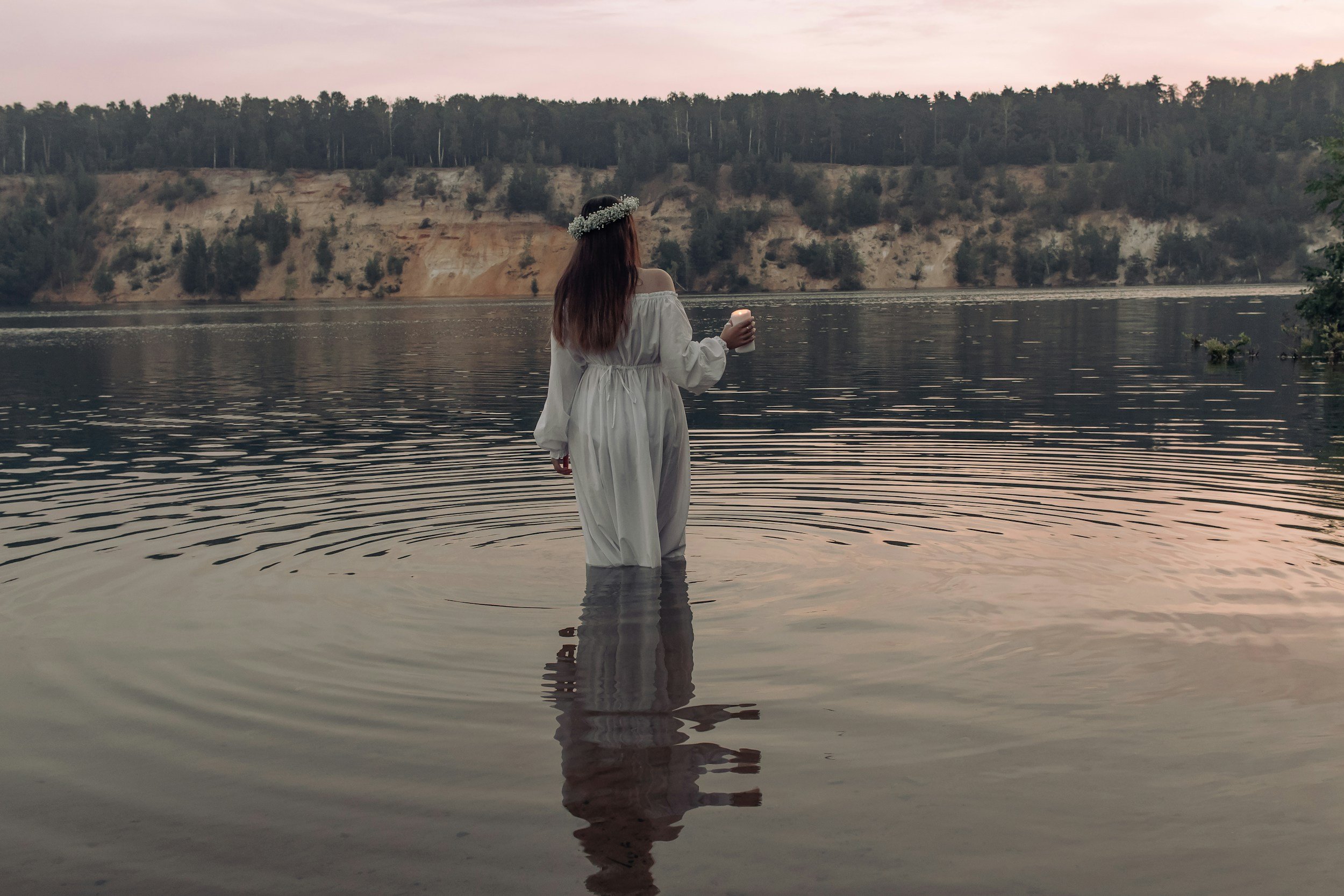 A wiccan in a white dress and flower crown standing in a calm body of water at sunset, holding a cup.