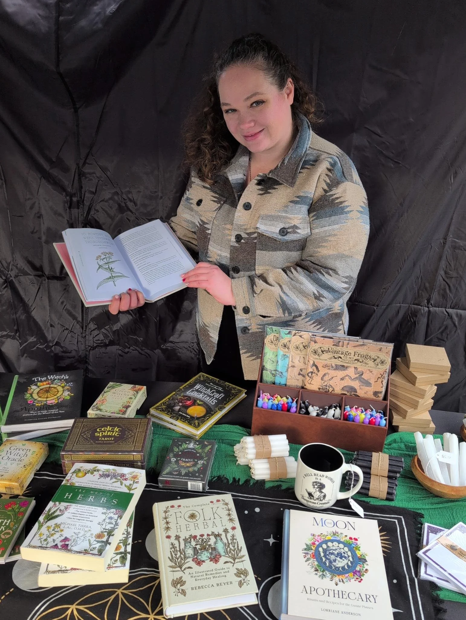 The apothecary holding a book on folk herbalism and welcoming you into her shop. On the table is witchy books and chime candles.
