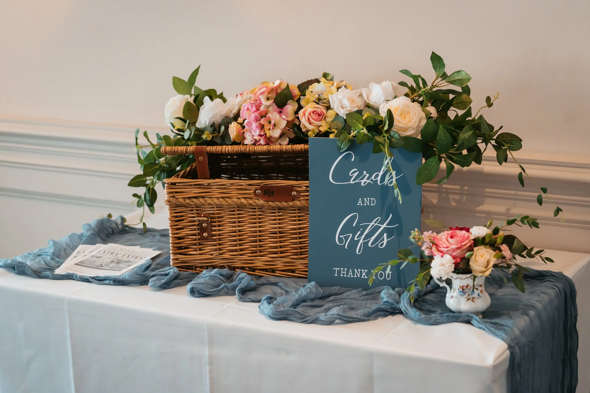 Wedding or event table with a wicker basket overflowing with pink and white flowers, a blue sign that reads 'Cards and Gifts Thank You', a small bouquet in a porcelain vase, and a newspaper on a white tablecloth with a blue fabric runner.
