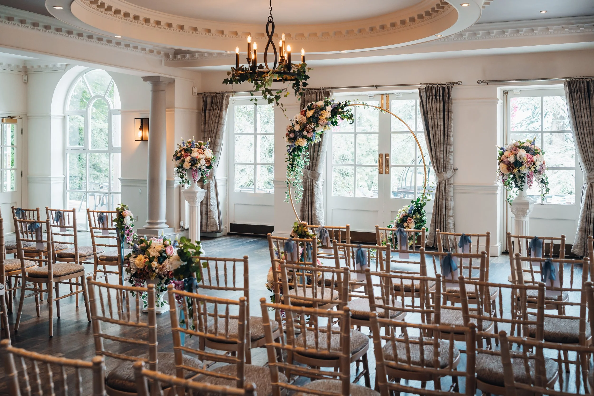 Wedding ceremony setup in a bright room with large windows, floral arrangements, a circular floral arch, wooden chairs with blue ribbons, and a chandelier hanging from the ceiling.
