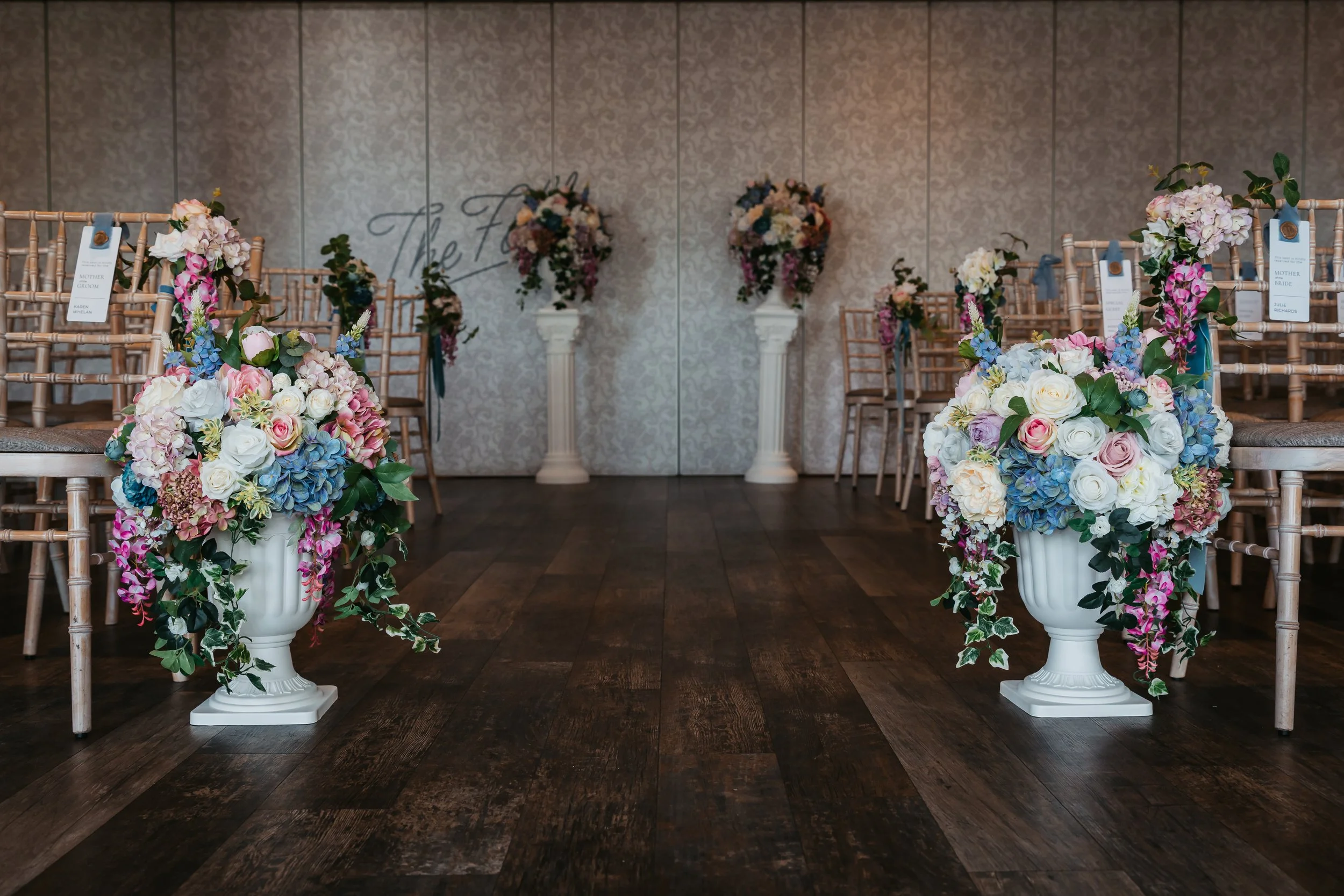 Elegant wedding setup with flower arrangements in white urns lining the aisle, pink, white, and blue hydrangeas, roses, and greenery, with chairs on either side and two pedestals with flowers at the end of the aisle.