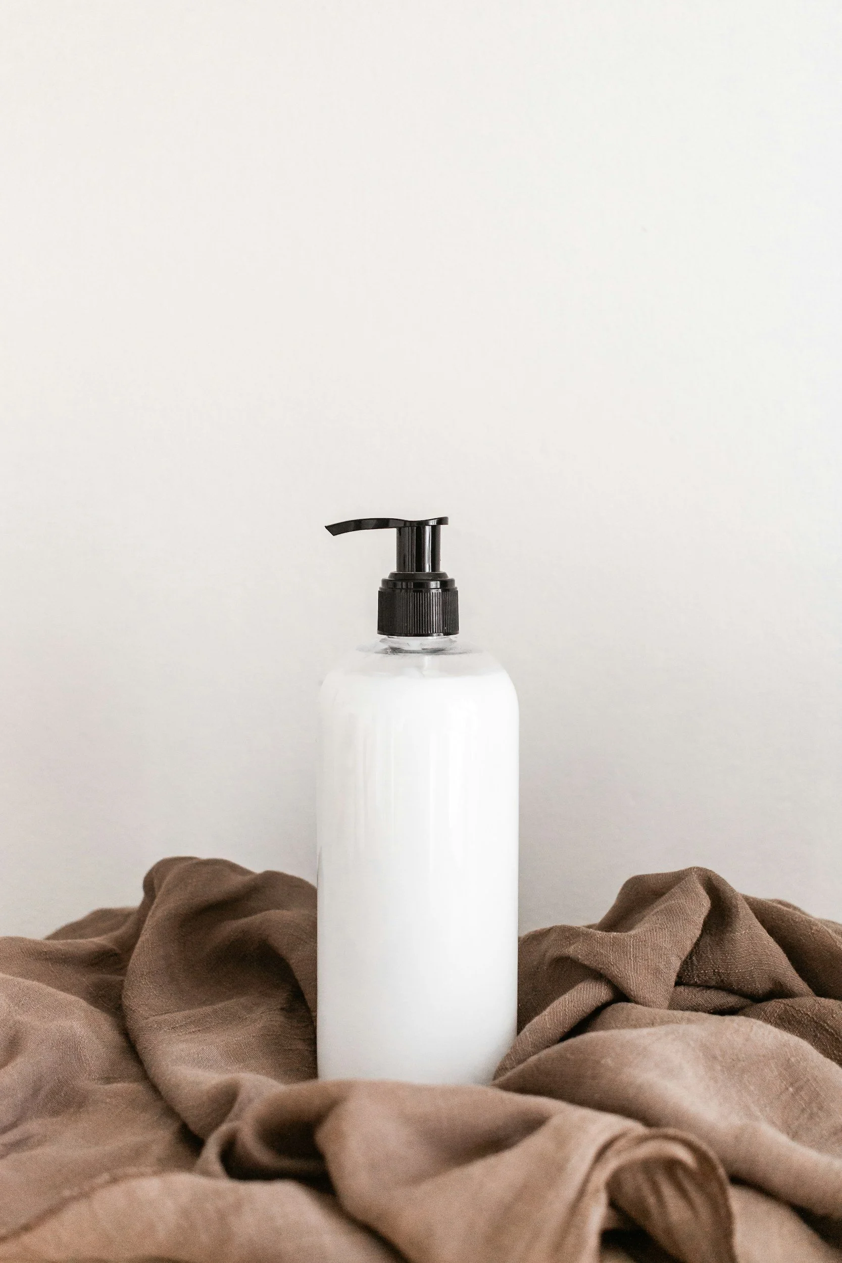 A white bottle with a black pump dispenser resting on a crumpled brown cloth against a plain white wall.