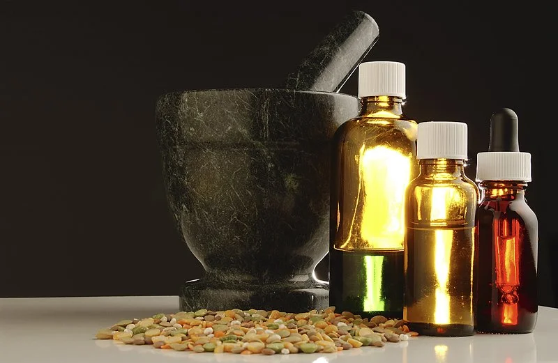 A black marble mortar and pestle with colorful pebble ingredients, and three small amber glass bottles with white caps and dropper tops.