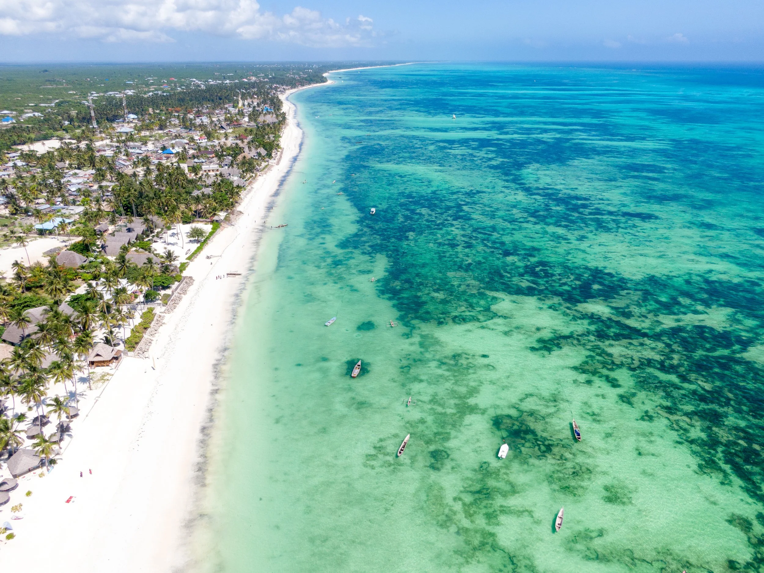 An aerial view of a tropical beach with white sand, palm trees, and resorts on land, alongside clear turquoise water with boats and ocean floor visible.