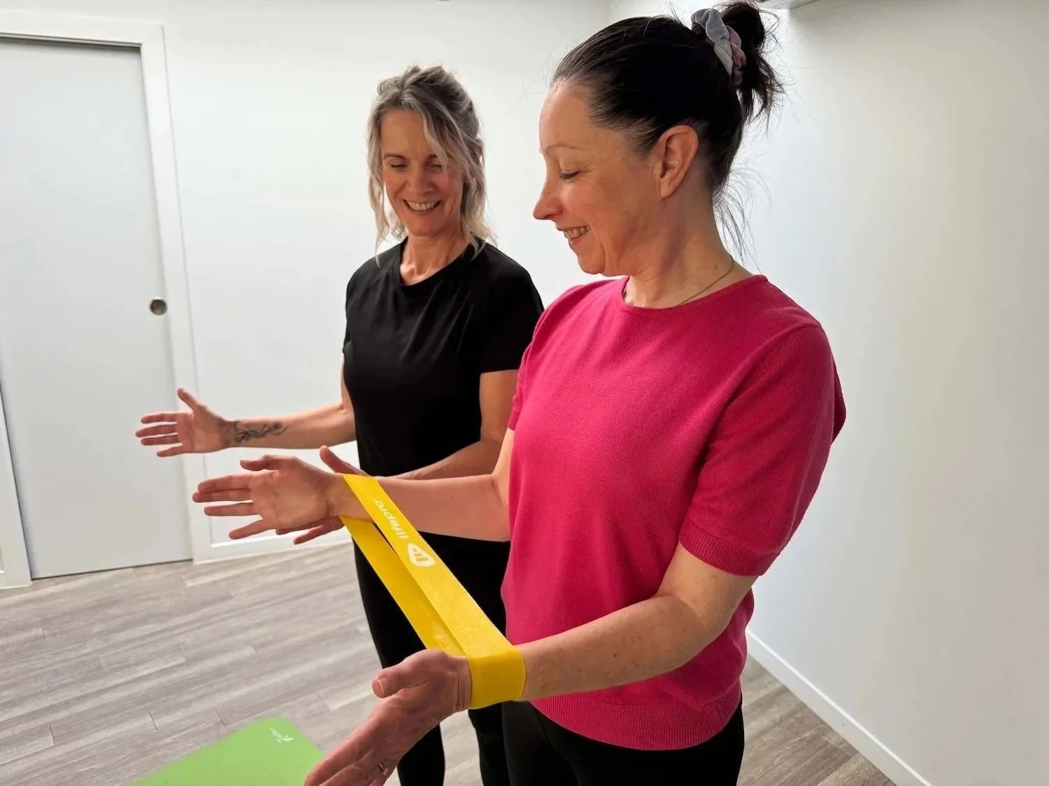 Two women standing indoors, one wearing a pink shirt and a yellow resistance band around her wrist, the other in black, smiling and engaging in a fitness activity.