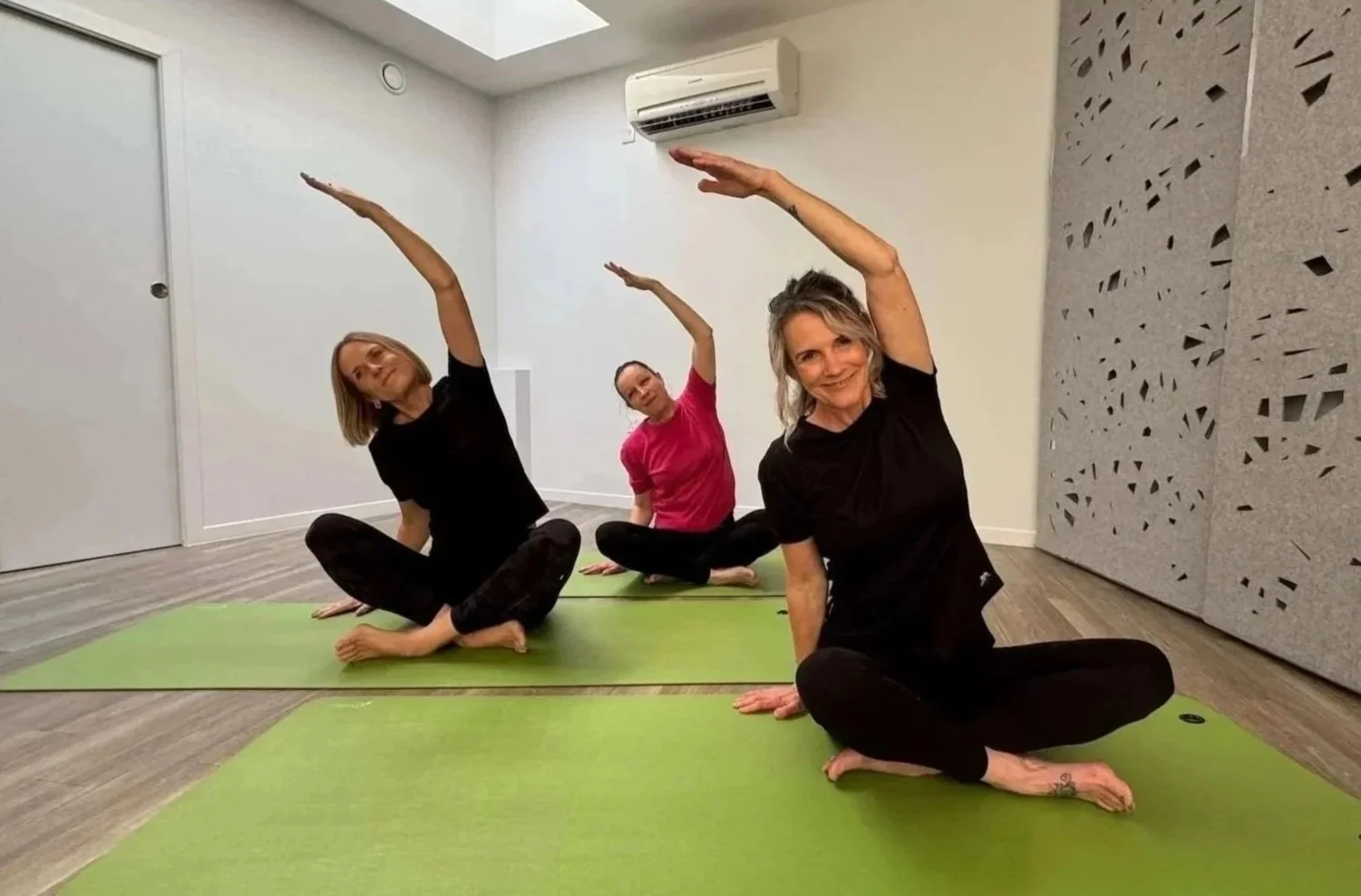 Three women practicing yoga on green mats in a room, sitting cross-legged and reaching overhead with one arm, smiling at the camera.