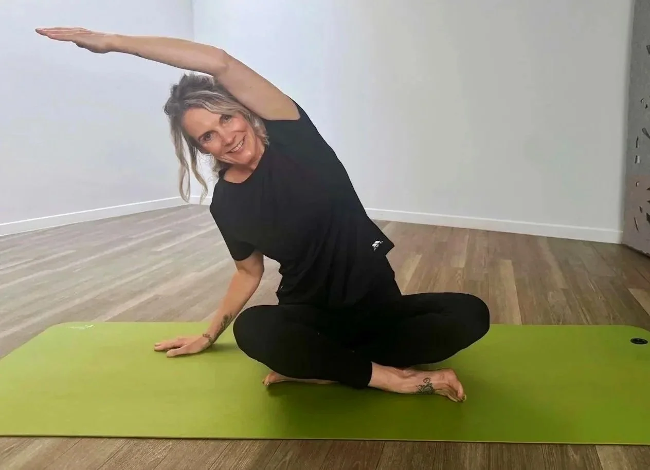A woman with gray hair in black athletic clothing practicing yoga on a green mat in a room with wooden floors, in a seated side bend pose with one arm extended over her head.