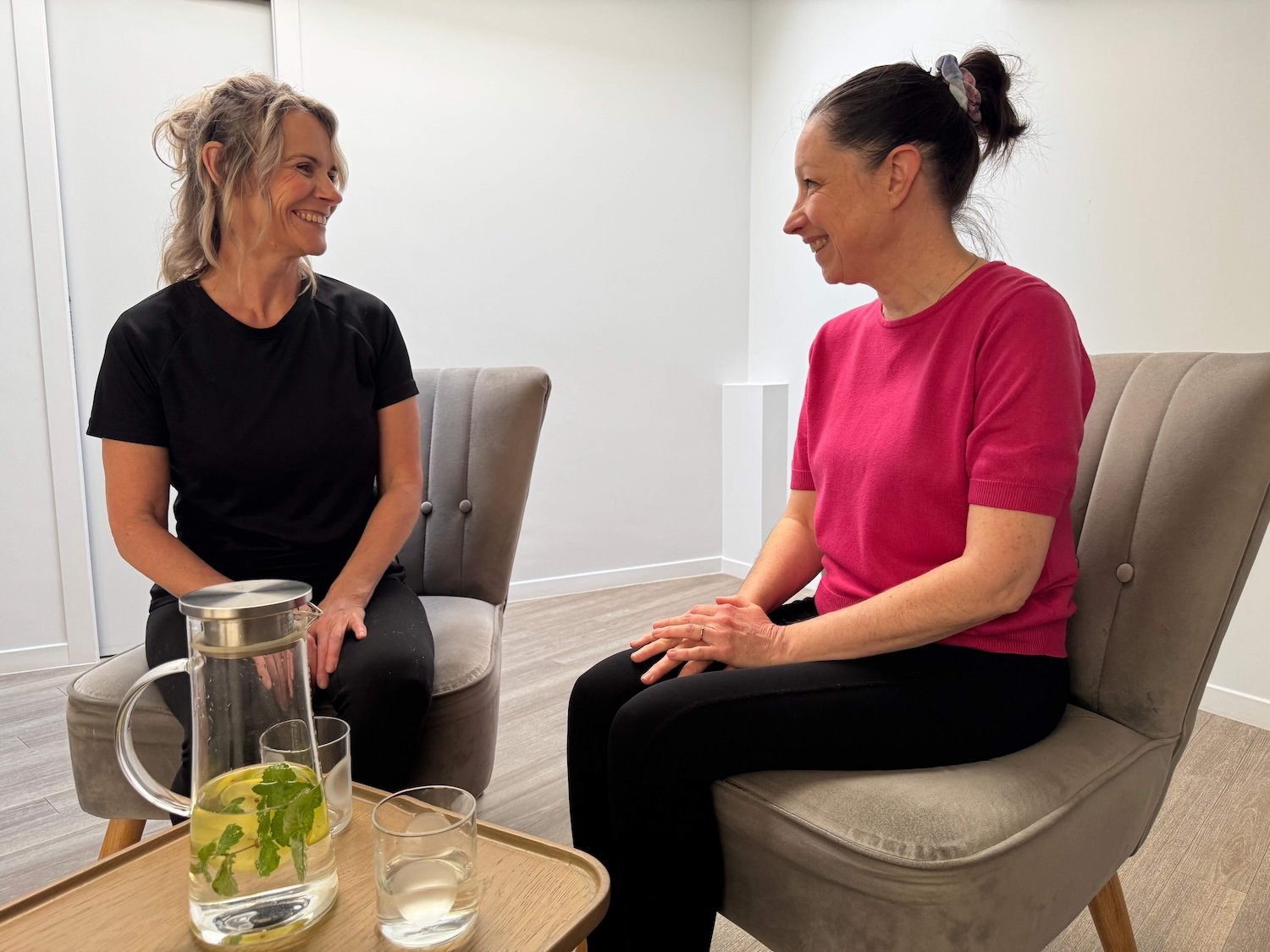 Two women sitting in chairs facing each other, smiling and engaged in conversation. A wooden tray with a glass pitcher of water, infused with mint and lemon, and two glasses is on a table in front of them. The setting appears to be a casual, indoor space.