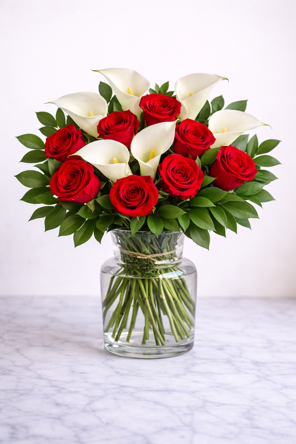 Bouquet of red roses and white calla lilies in a glass vase on a white marble surface.