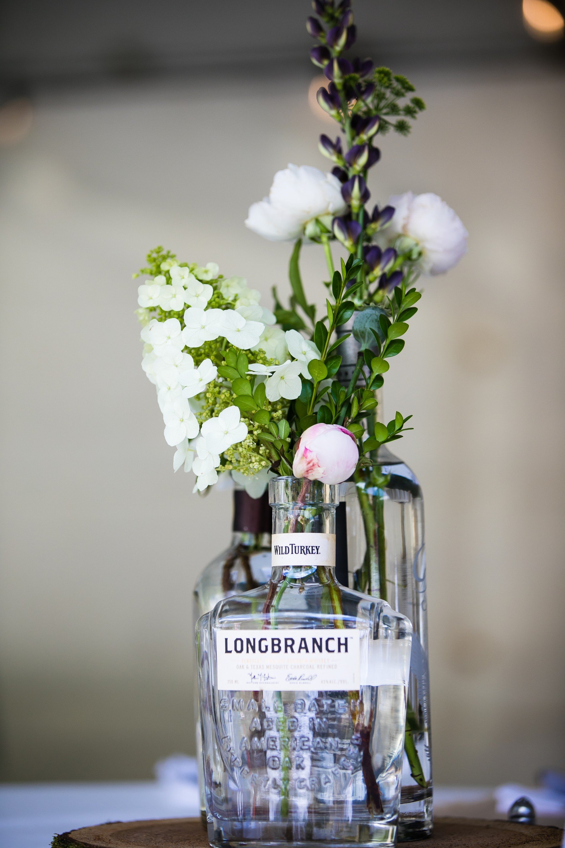 A glass bottle with flowers inside, including white hydrangeas, a pink peony, and purple and white flowers, placed on a wooden surface.