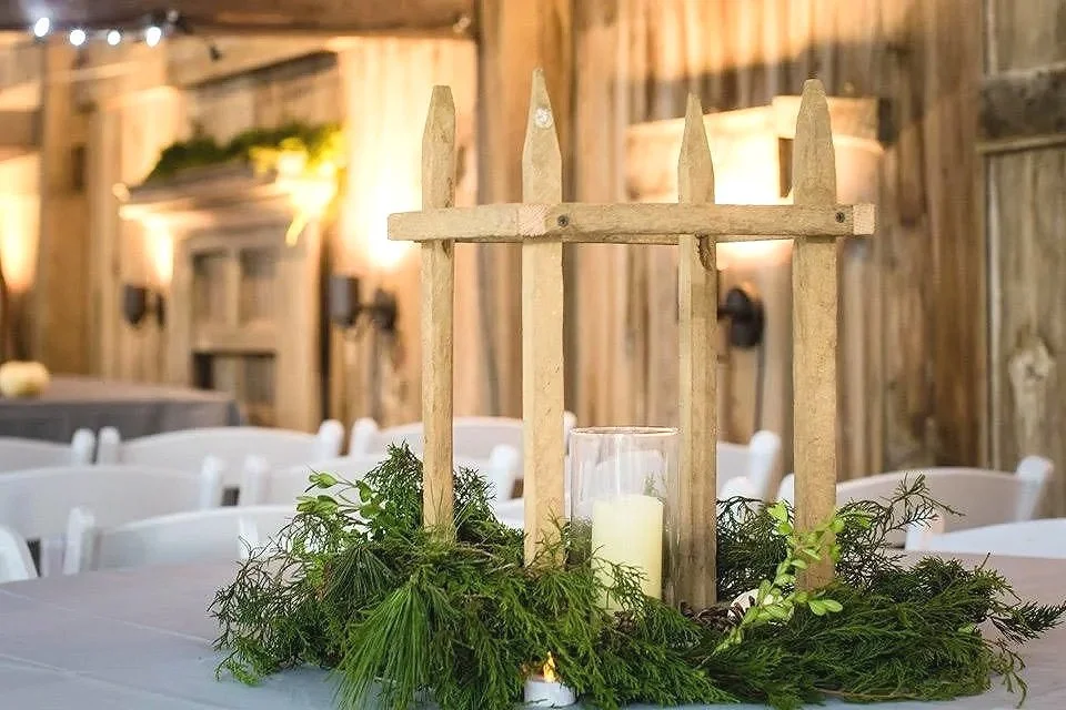 Rustic wedding centerpiece with a wooden spiked fence, green foliage, and a white candle in a glass holder on a decorated table in a wooden barn setting.